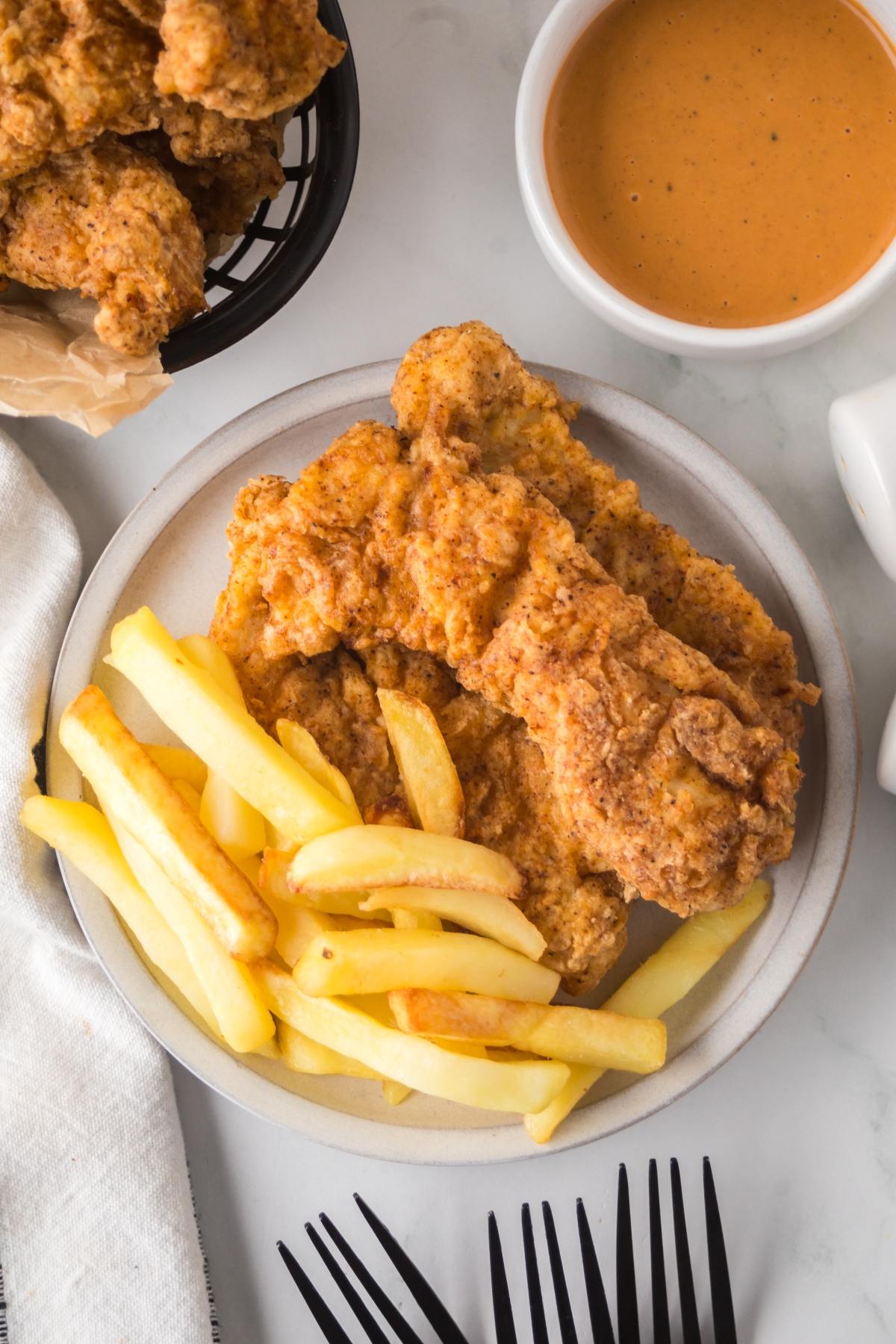 Plate of fried chicken tenders and French fries, with dipping sauce in a bowl nearby.
