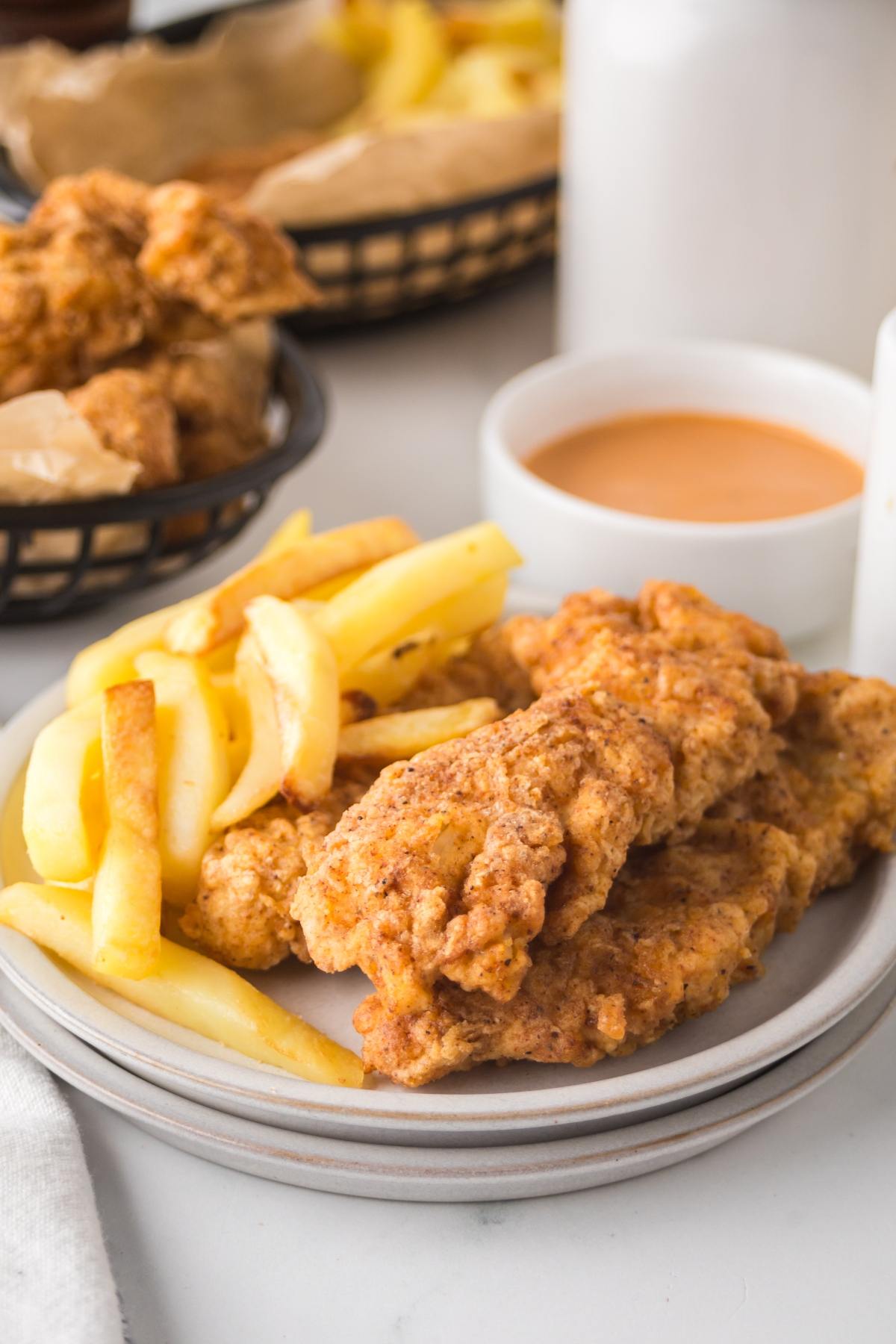 Plate of fried chicken tenders and French fries with dipping sauce and more food in the background.