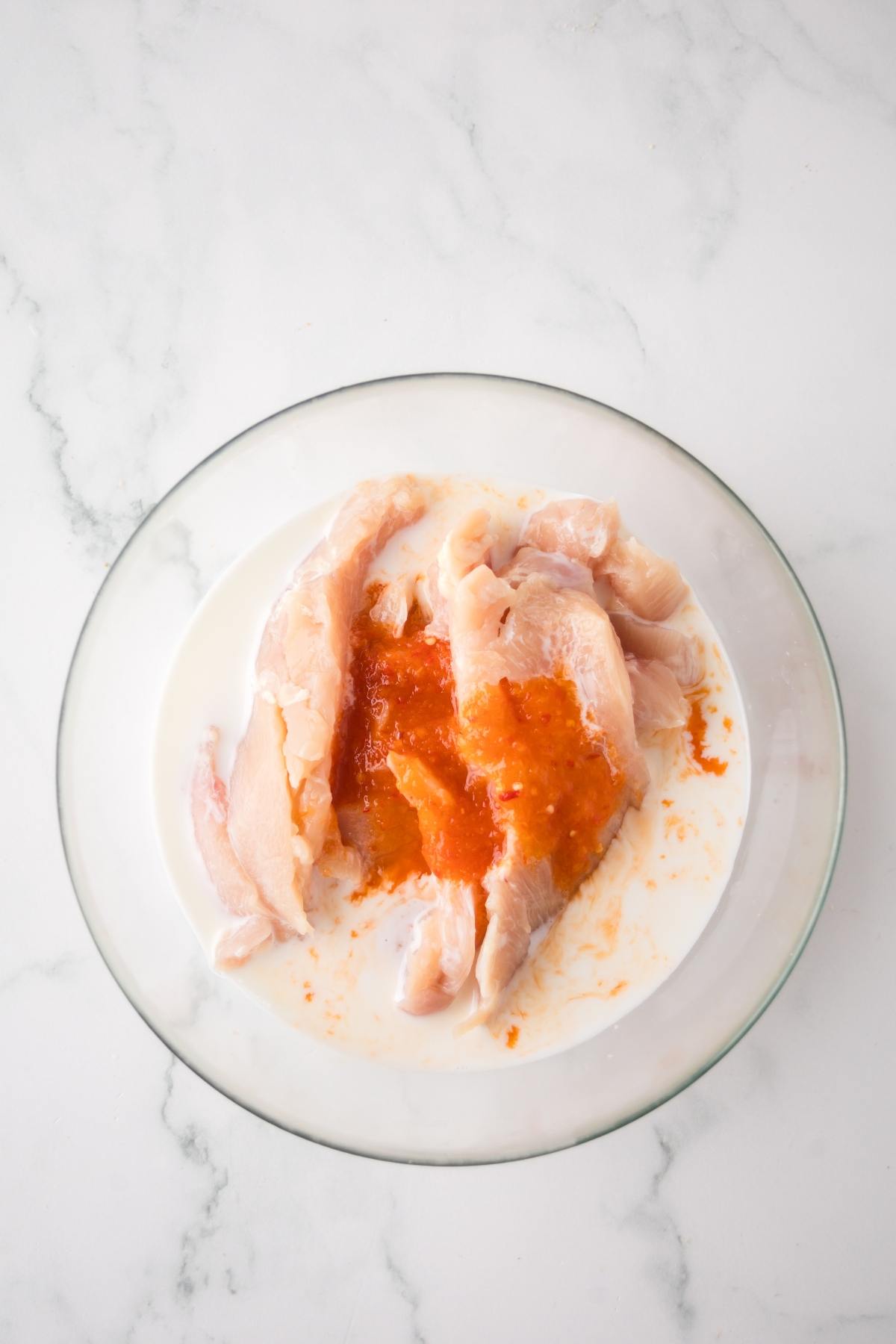 Raw chicken breasts in a bowl with white marinade and orange sauce on a marble countertop.