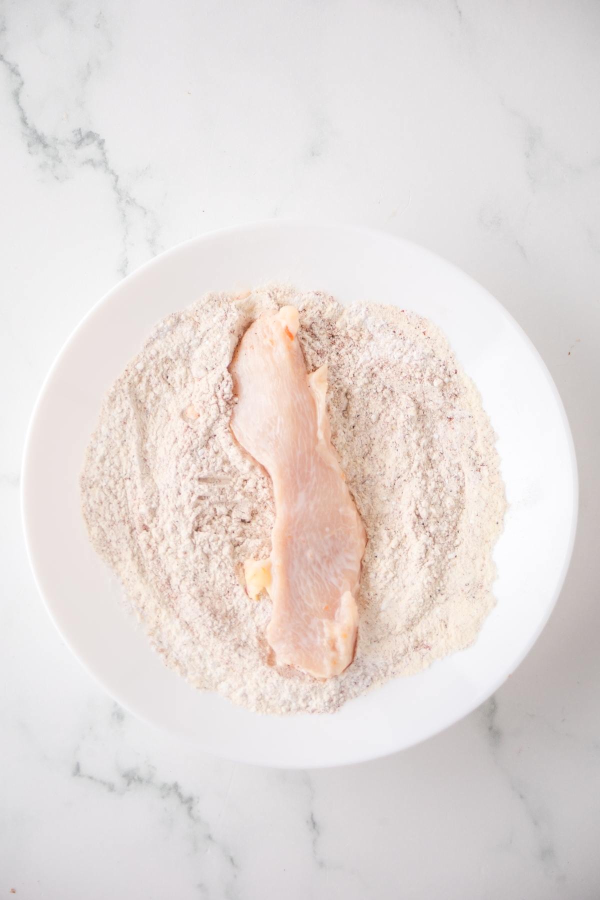 Raw chicken breast being coated in flour mixture on a white plate, on a white marble surface.