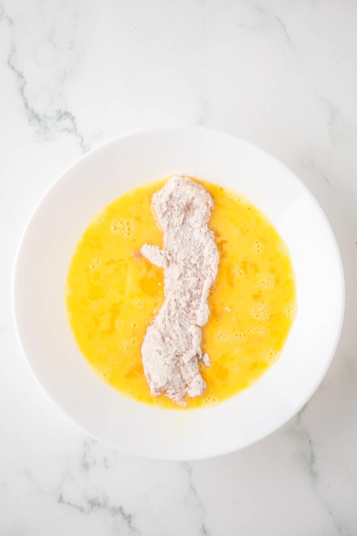 A breaded chicken strip dipped in beaten egg in a white bowl on a marble surface.