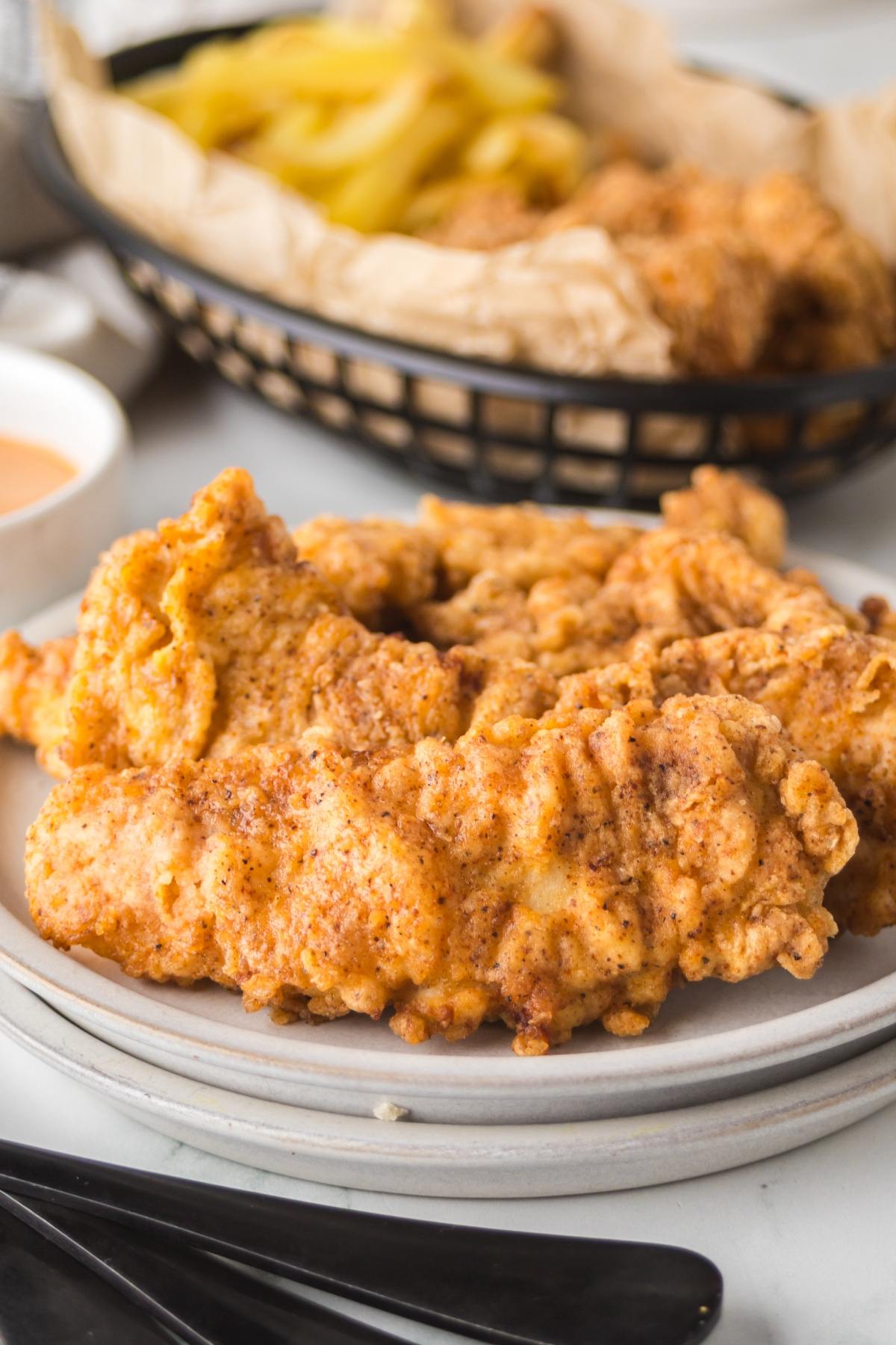 Crispy fried chicken tenders on a plate with fries in a basket in the background.