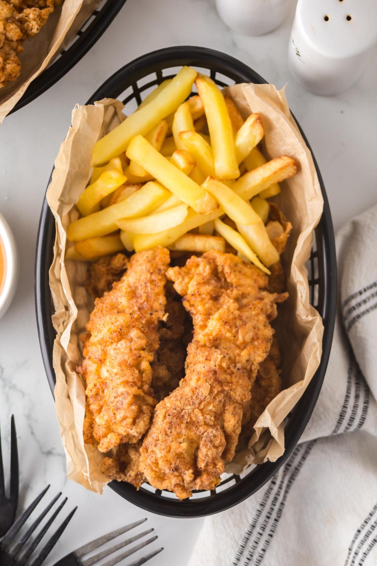 Basket of crispy fried chicken tenders and golden French fries on parchment paper, with a fork nearby.
