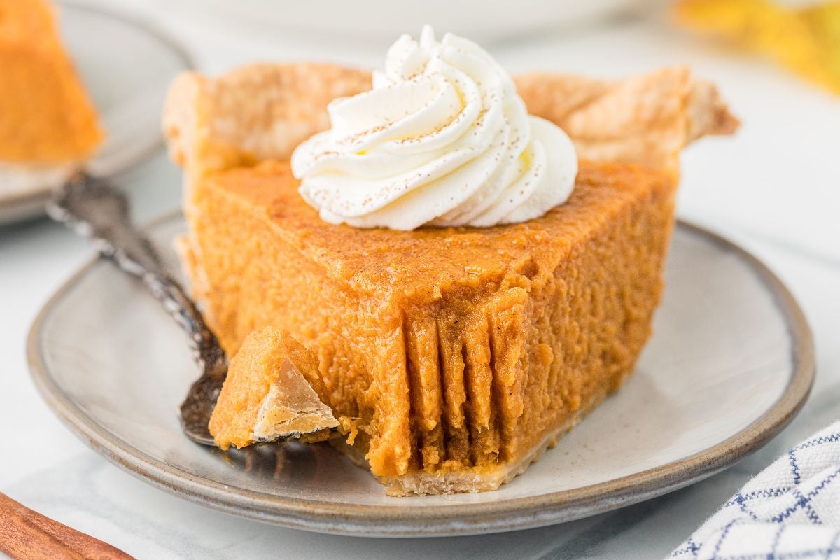 A slice of sweet potato pie with a dollop of whipped cream on top, on a plate with a fork and a bite taken out.