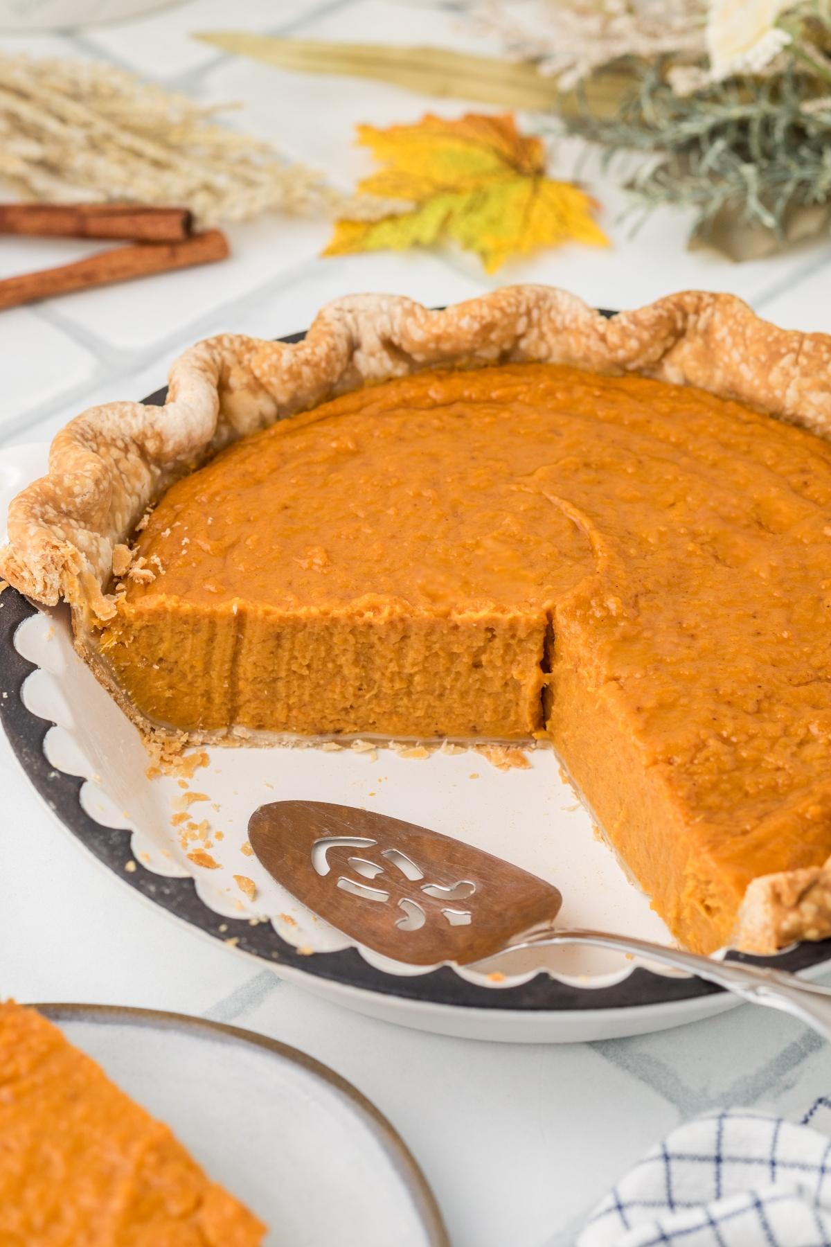 Pumpkin pie with a slice missing, showing creamy filling and flaky crust, on a white table with fall decor.