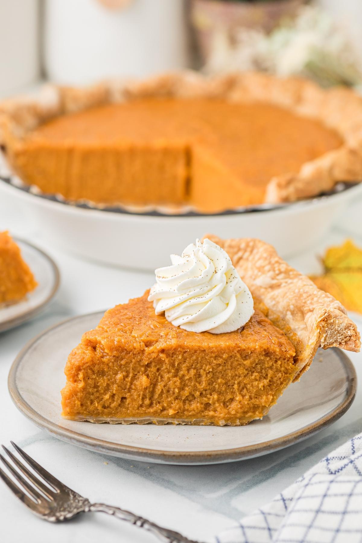 A slice of pumpkin pie topped with whipped cream sits on a plate, with the whole pie in the background.