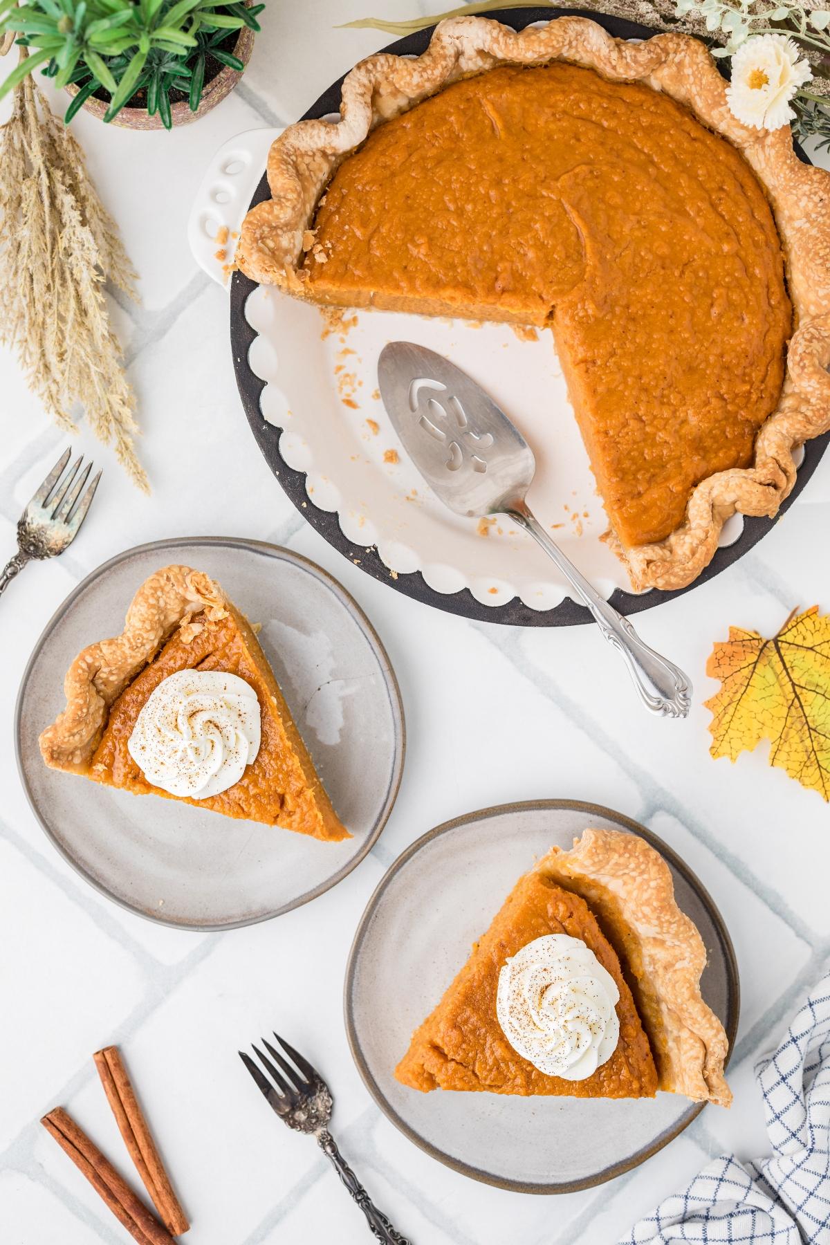 Pumpkin pie with two slices on plates, topped with whipped cream. Pie server and fall decor are nearby.
