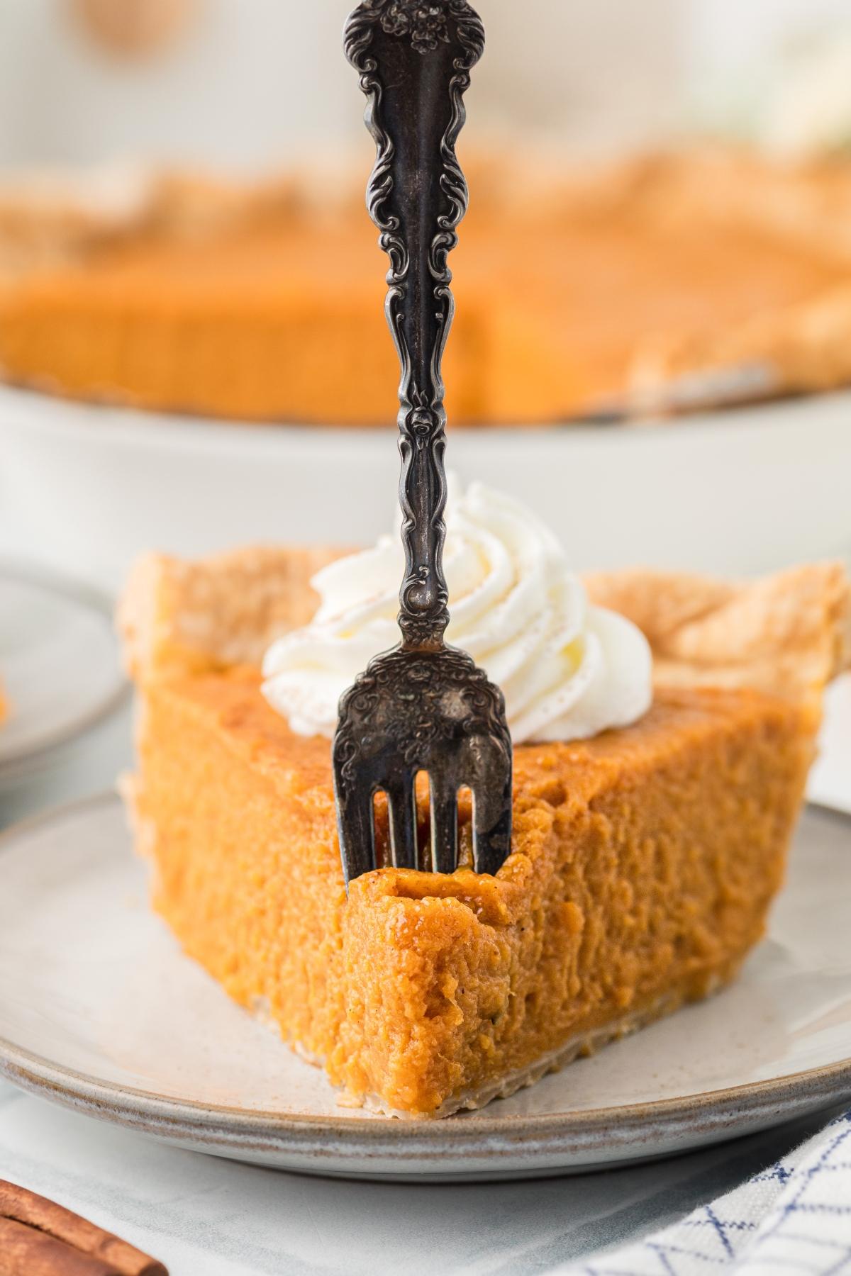 A slice of pumpkin pie with whipped cream, pierced by an ornate fork on a plate.