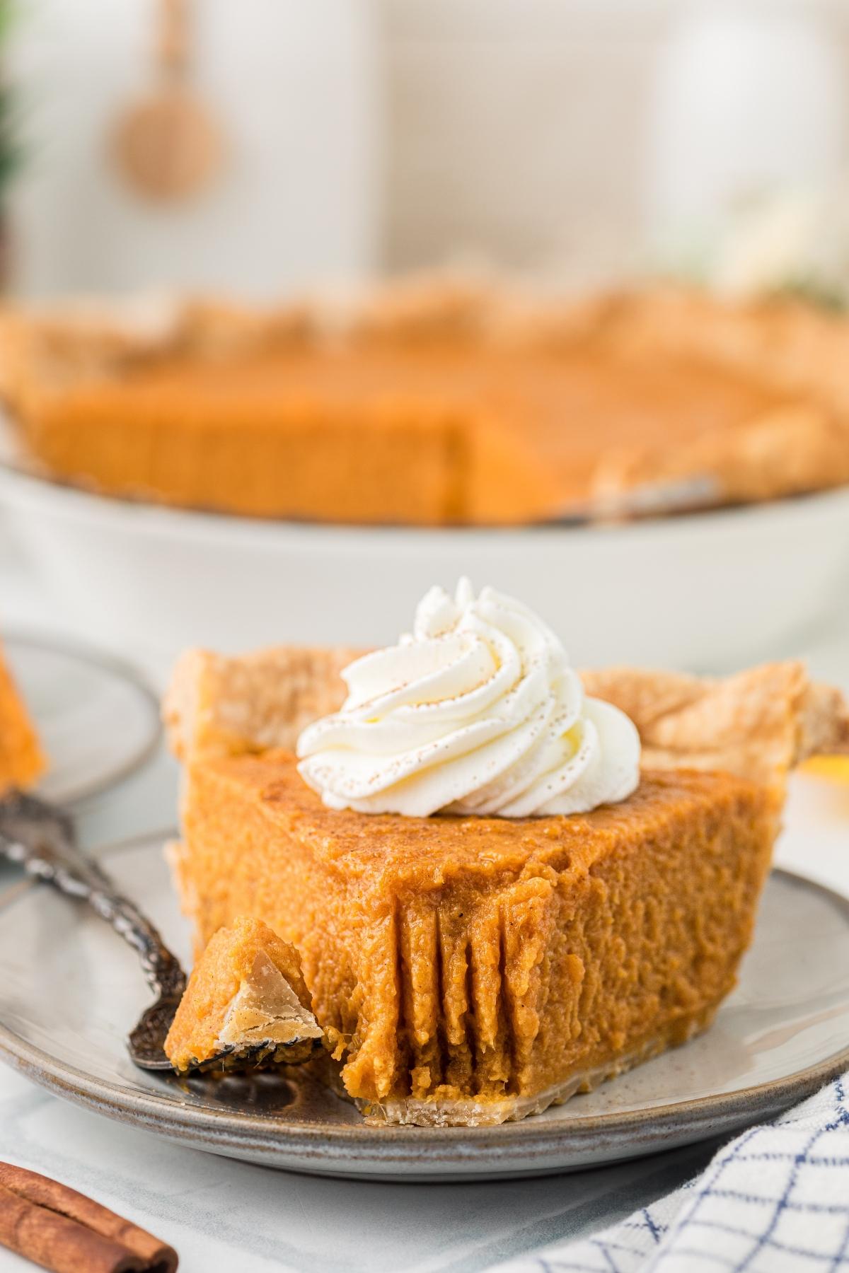 A slice of pumpkin pie with whipped cream on top, served on a plate with a fork and a bite taken out.