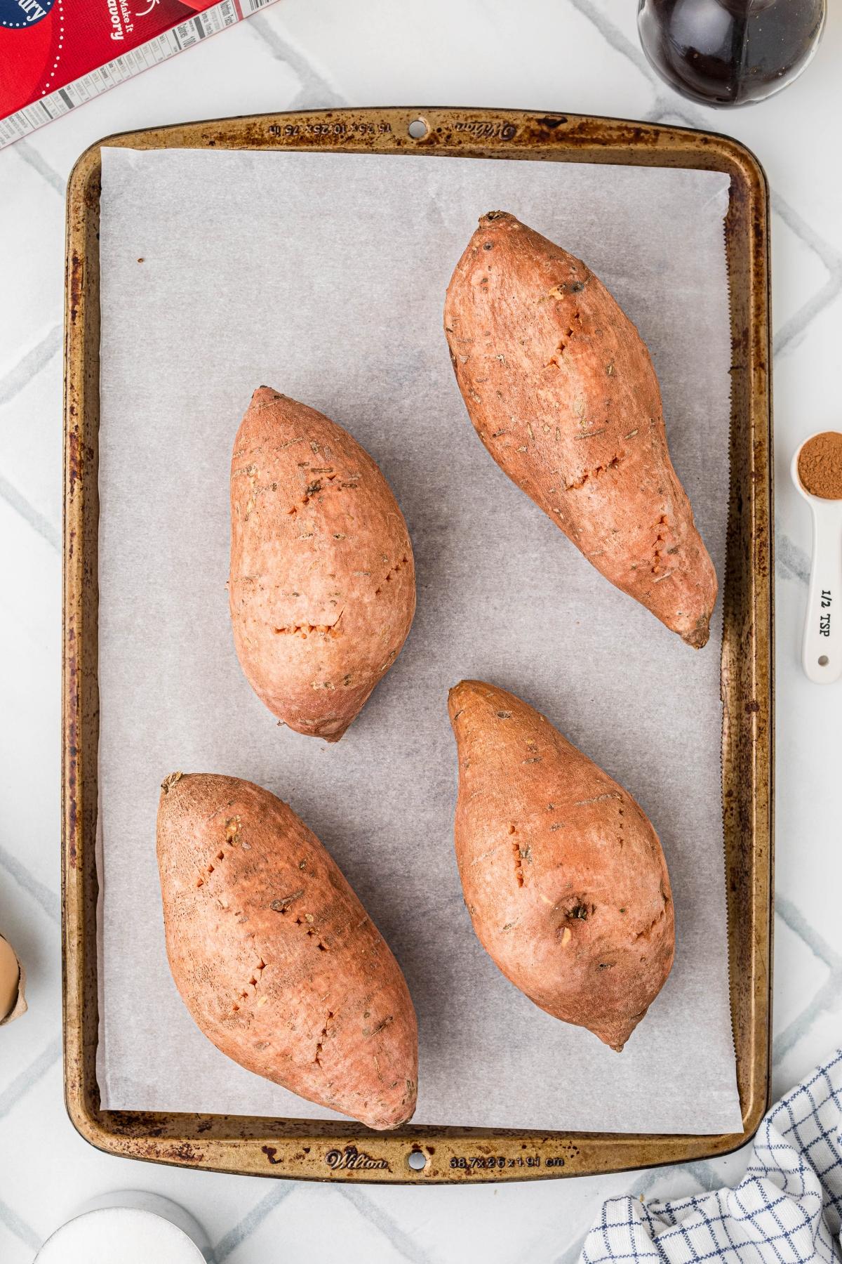 Four whole sweet potatoes on a parchment-lined baking sheet, ready to be baked.