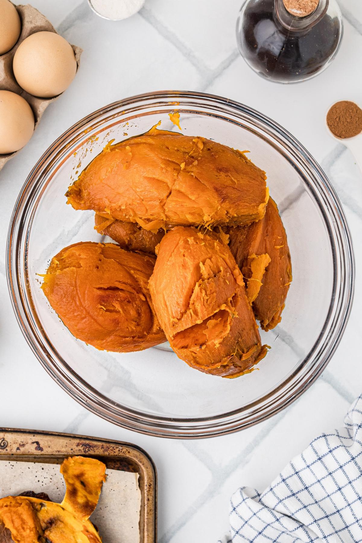 A glass bowl filled with roasted pumpkin flesh, surrounded by eggs, spices, and a baking tray.