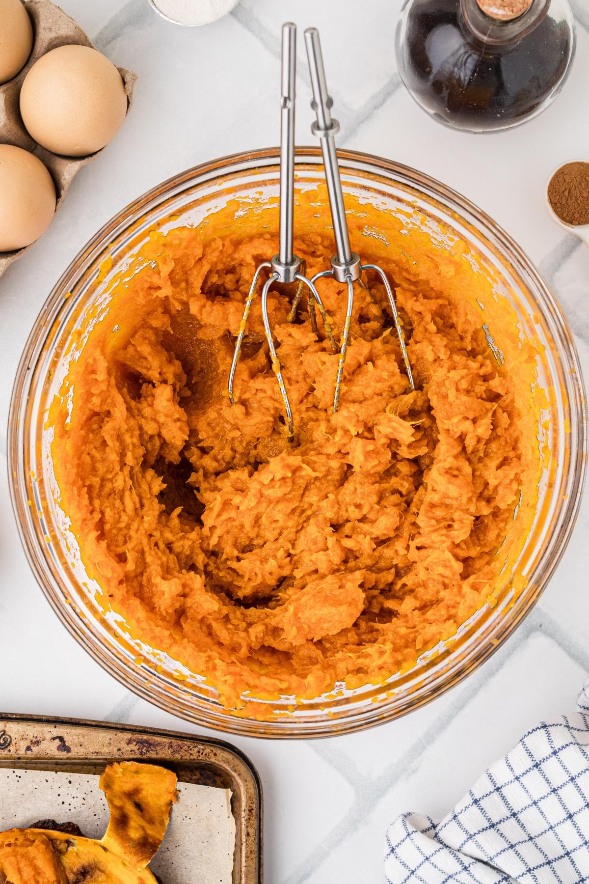 A glass bowl of mashed sweet potatoes with metal beaters, surrounded by eggs and kitchen items.