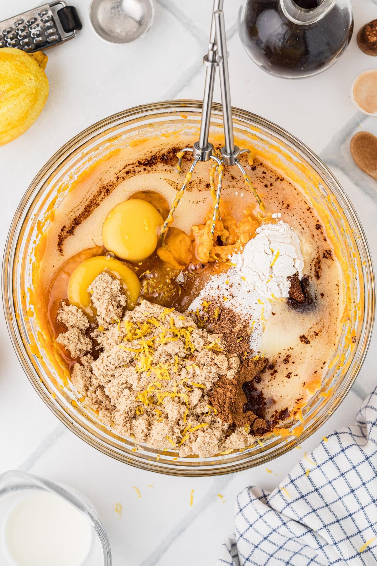 A mixing bowl with eggs, brown sugar, flour, spices, and lemon zest, ready to be blended with a hand mixer.
