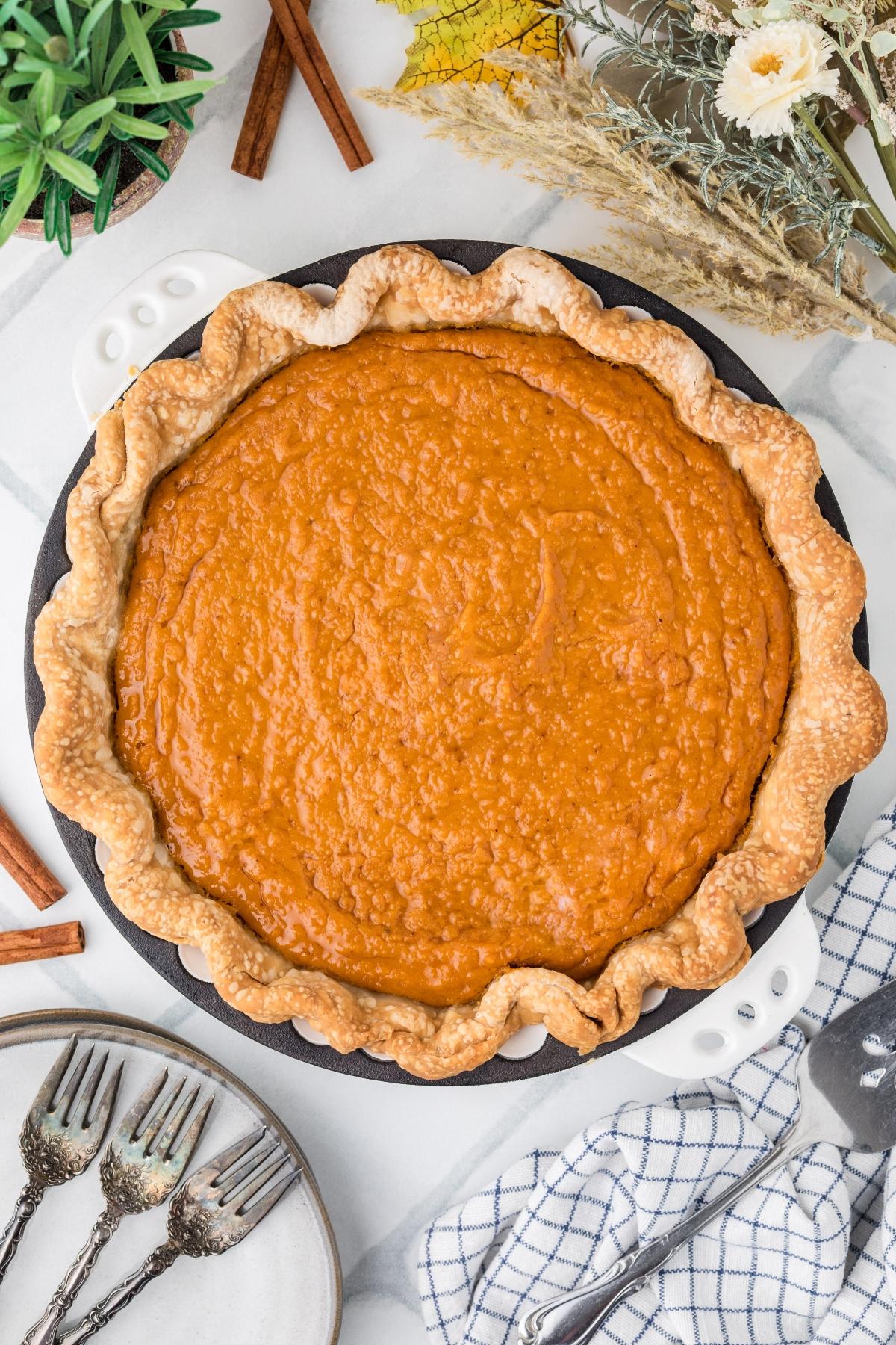 A freshly baked pumpkin pie with a golden crust on a marble surface, surrounded by utensils and autumn decor.
