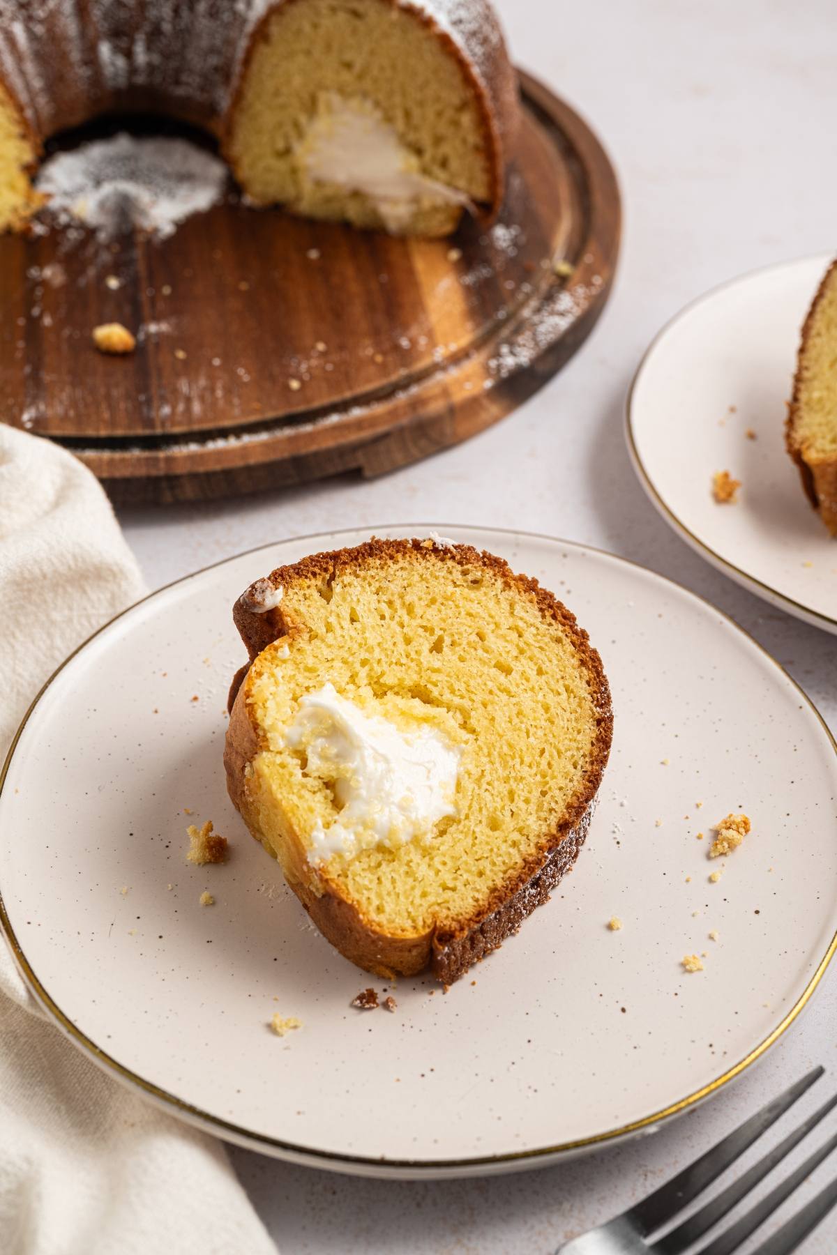 A slice of bundt cake with cream filling on a plate, with more cake in the background on a wooden board.