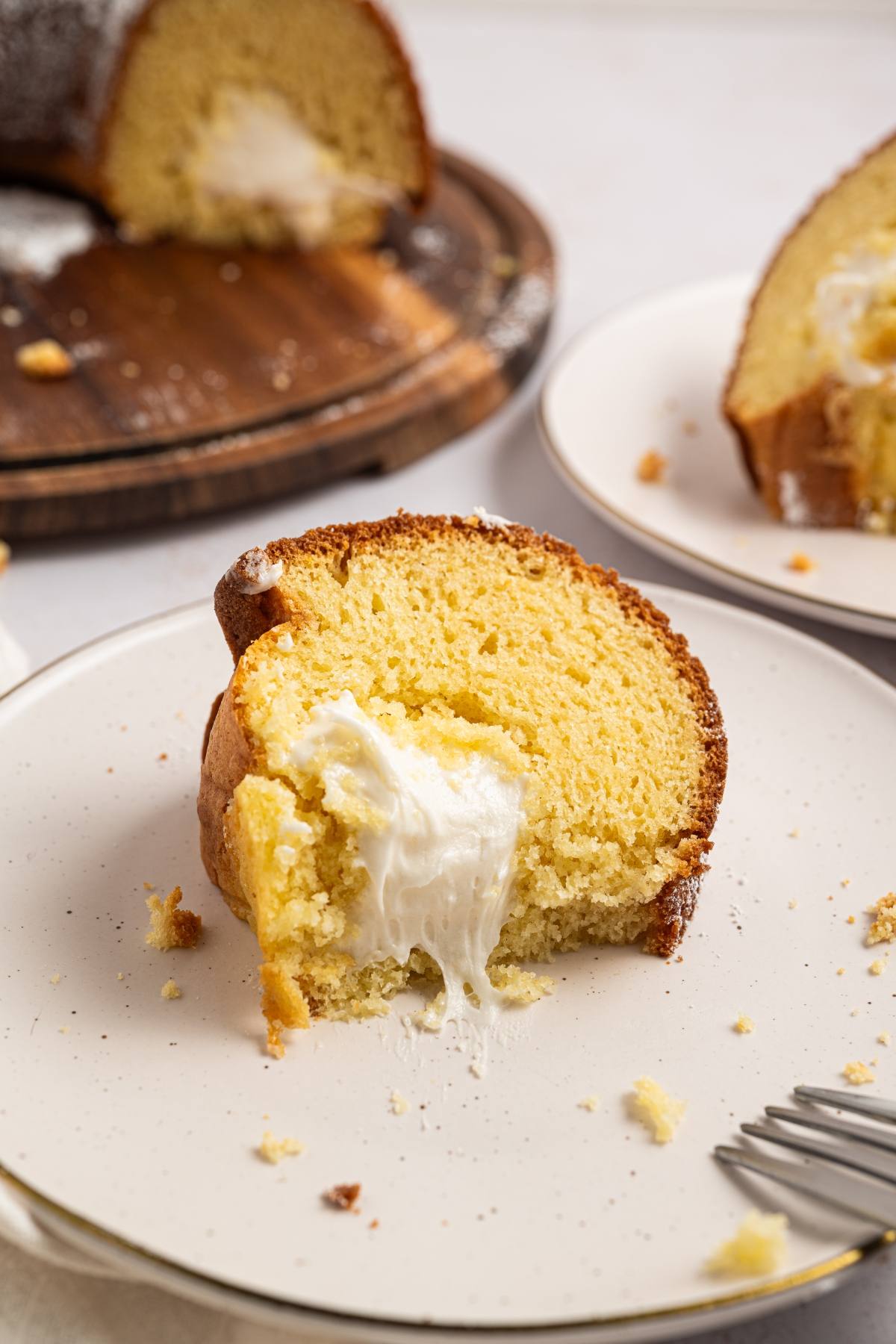 A slice of yellow cake with cream filling on a plate, with a whole cake and another slice in the background.
