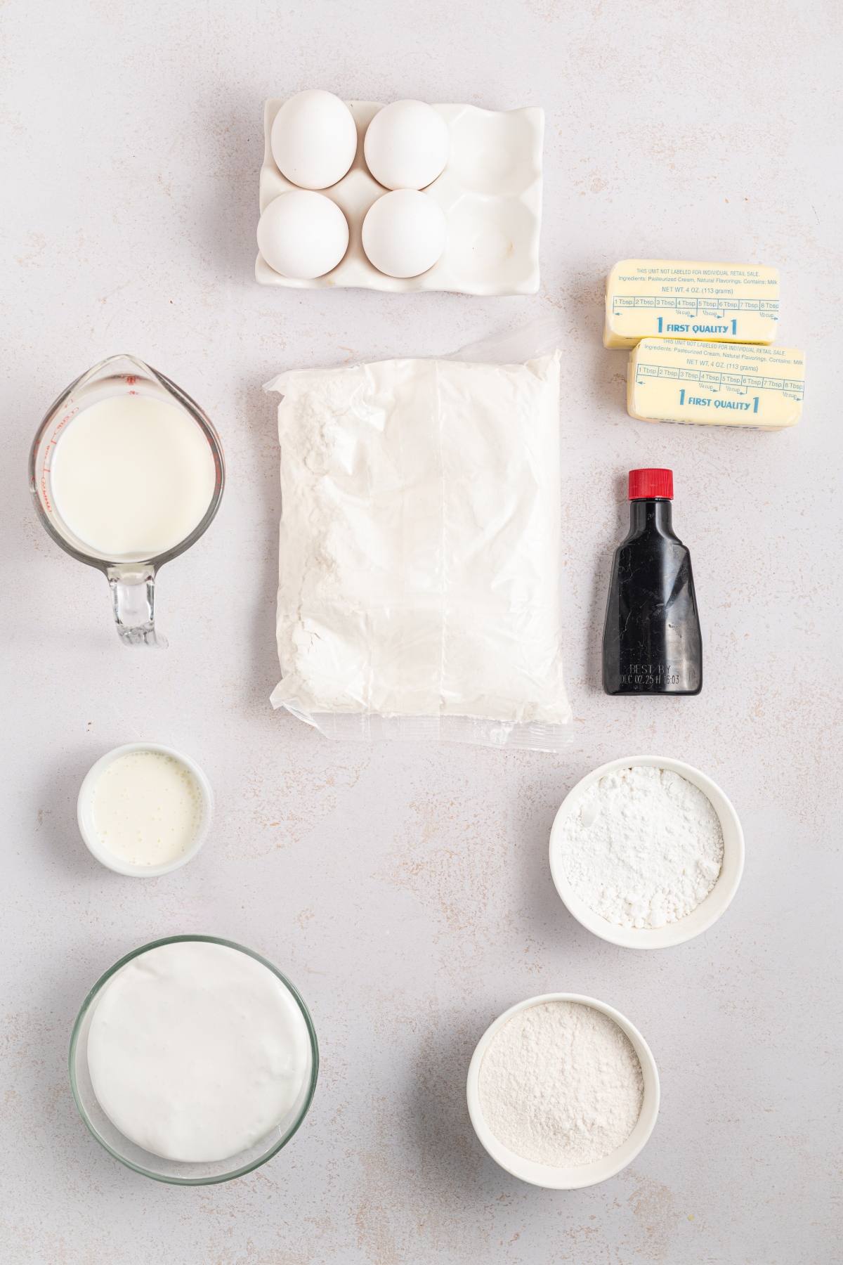 Baking ingredients arranged on a light surface: eggs, butter, flour, milk, vanilla, sugar, and cream.