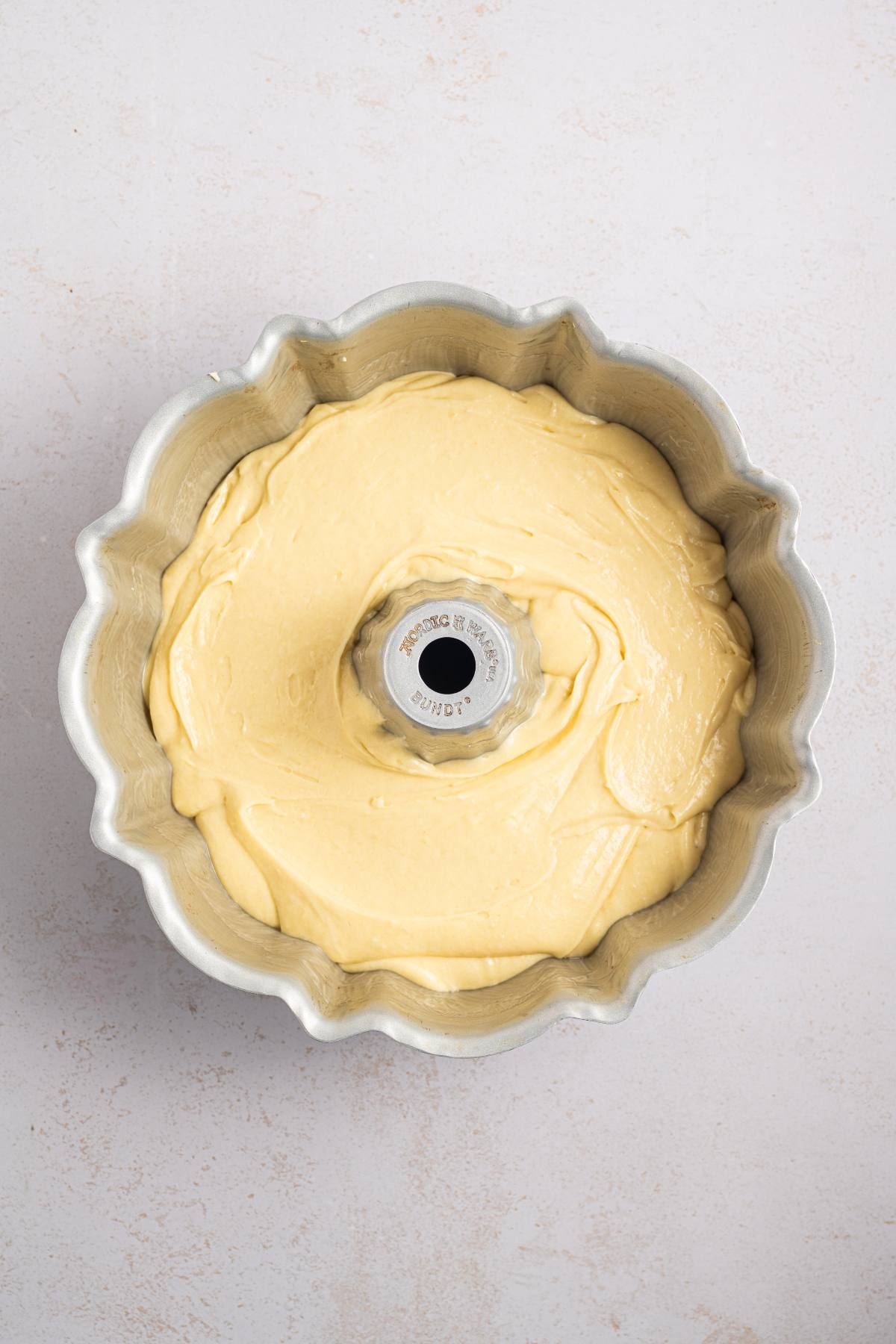 Cake batter evenly spread in a Bundt pan, ready to be baked, on a light-colored surface.