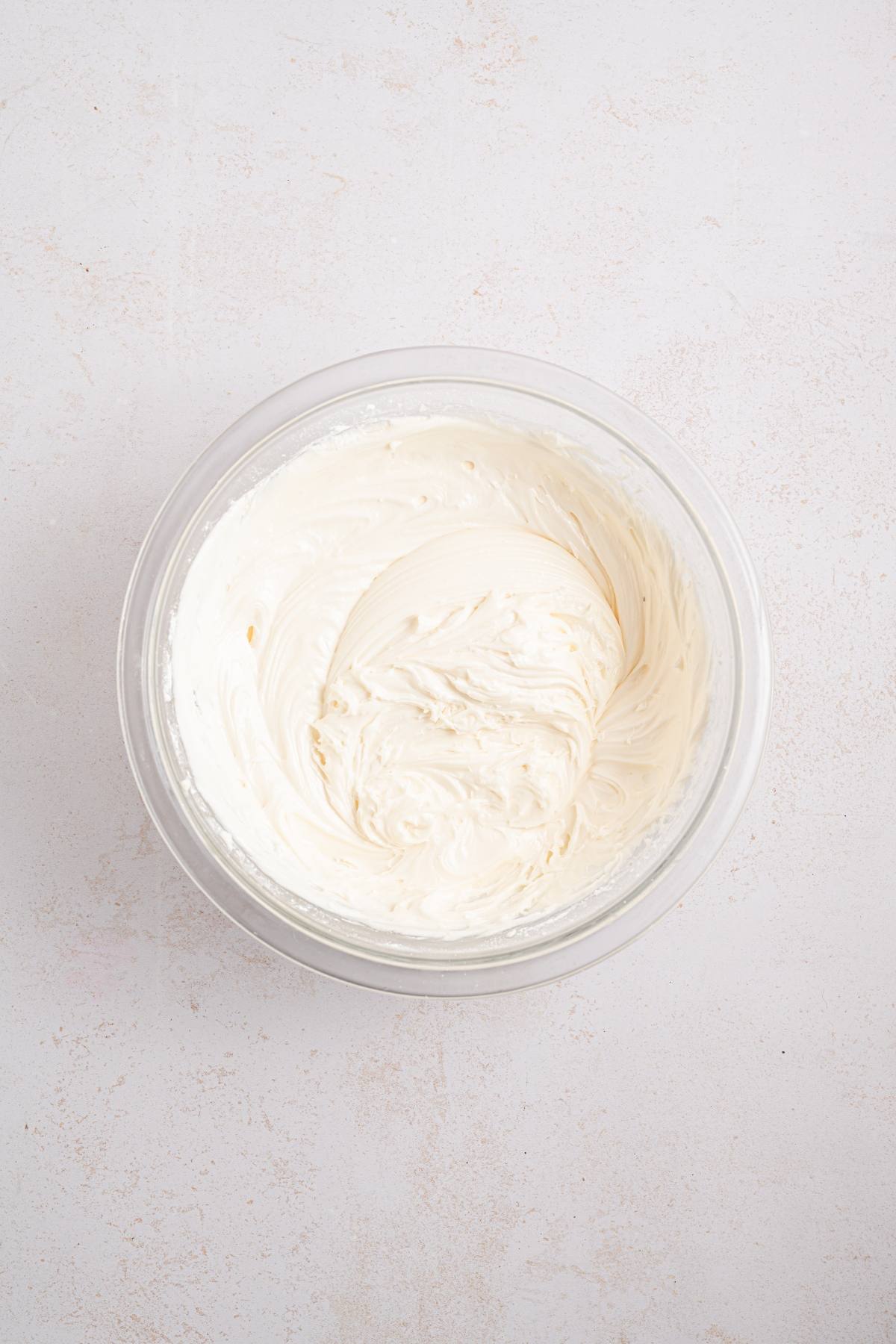 A clear bowl of whipped cream cheese frosting on a light, textured surface.