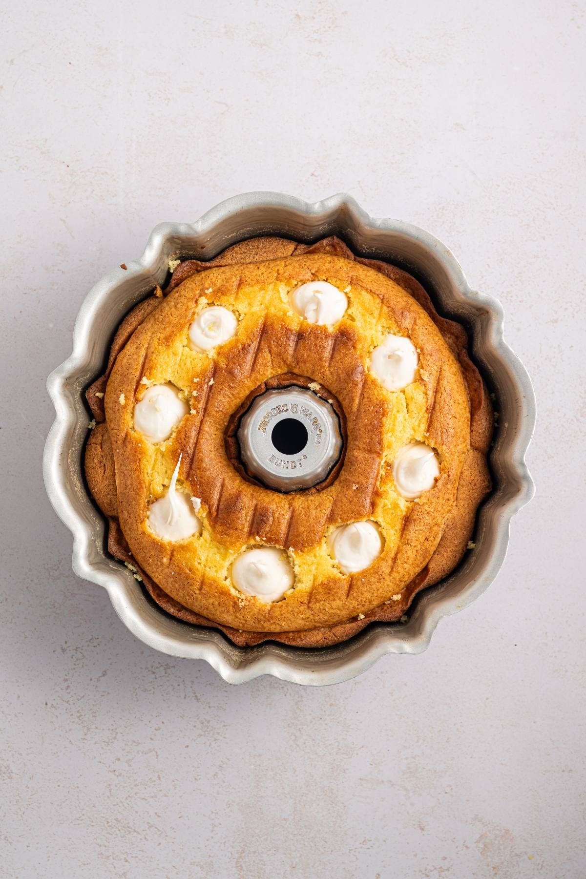 A bundt cake with white icing dots on top, in a metal bundt pan, on a light surface.