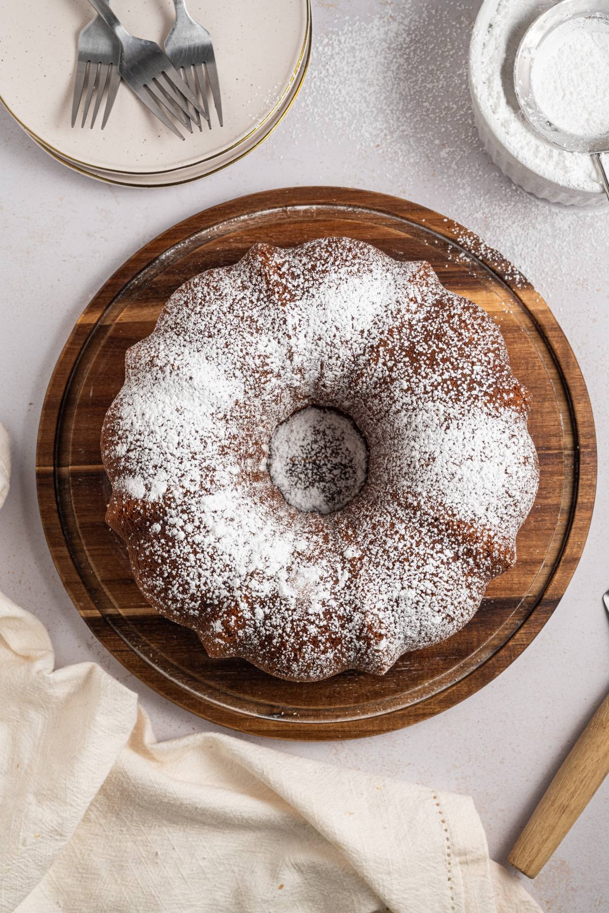 A Bundt cake dusted with powdered sugar on a wooden board, with plates, forks, and a napkin nearby.