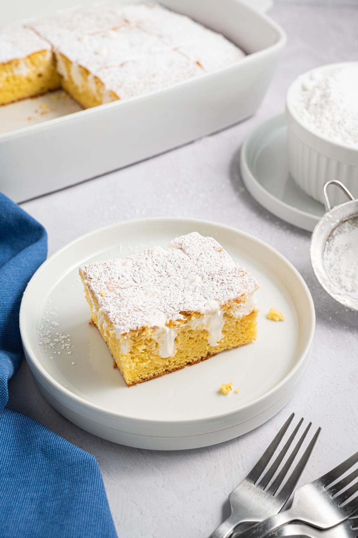 Square slice of yellow cake with powdered sugar and icing on a white plate, next to forks and a blue napkin.