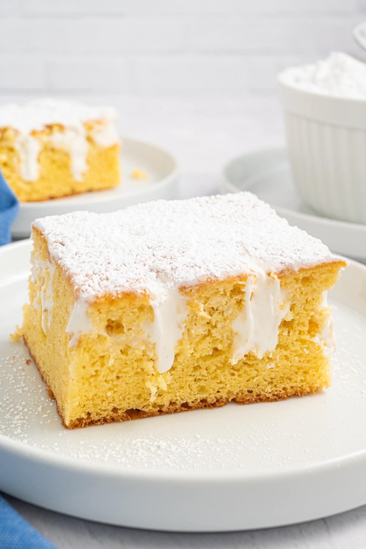 A square slice of yellow cake with white icing and powdered sugar on top, served on a white plate.