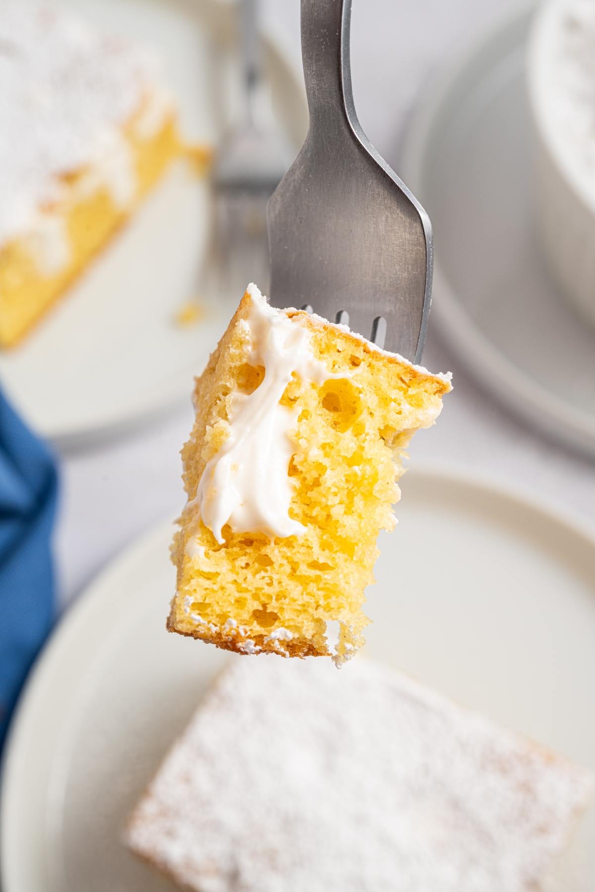 A fork holds a piece of yellow cake with white frosting, above a plate with more cake.