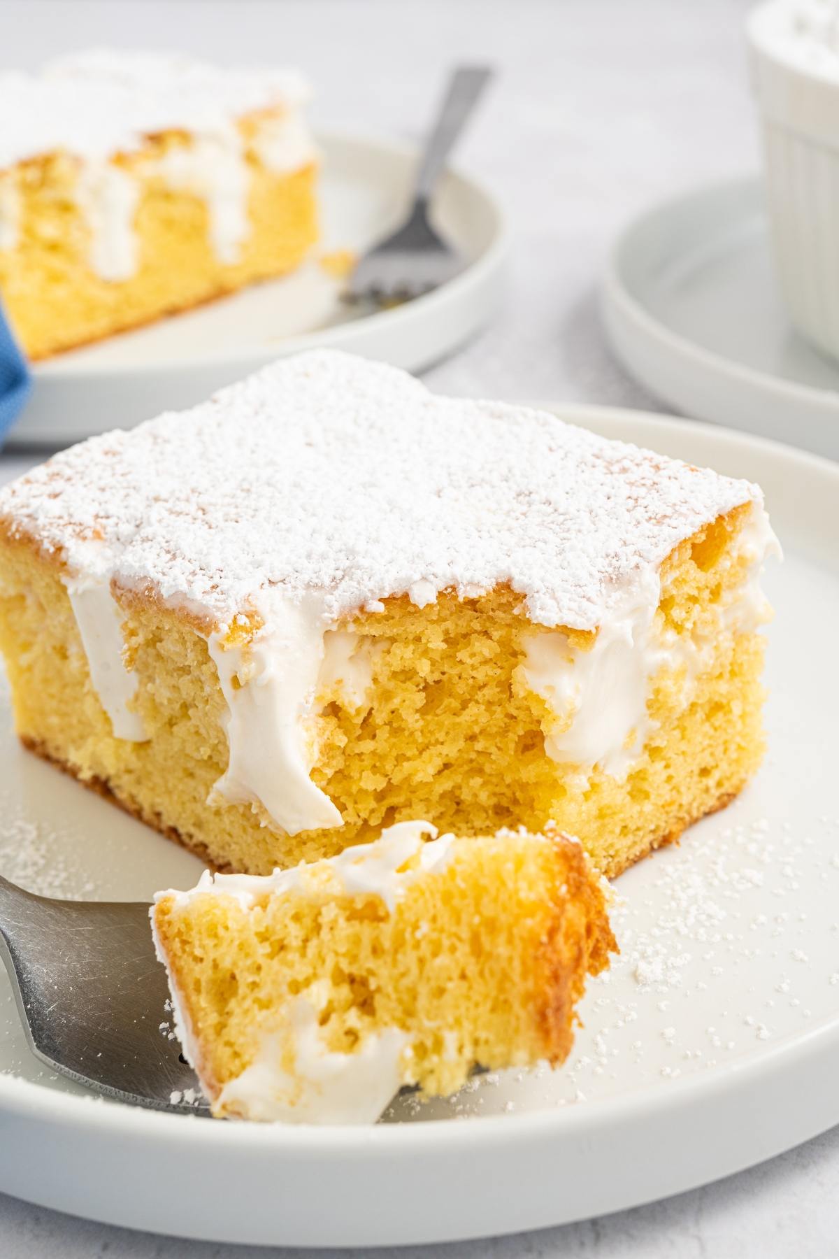 A slice of yellow cake with cream filling and powdered sugar on top, served on a white plate.