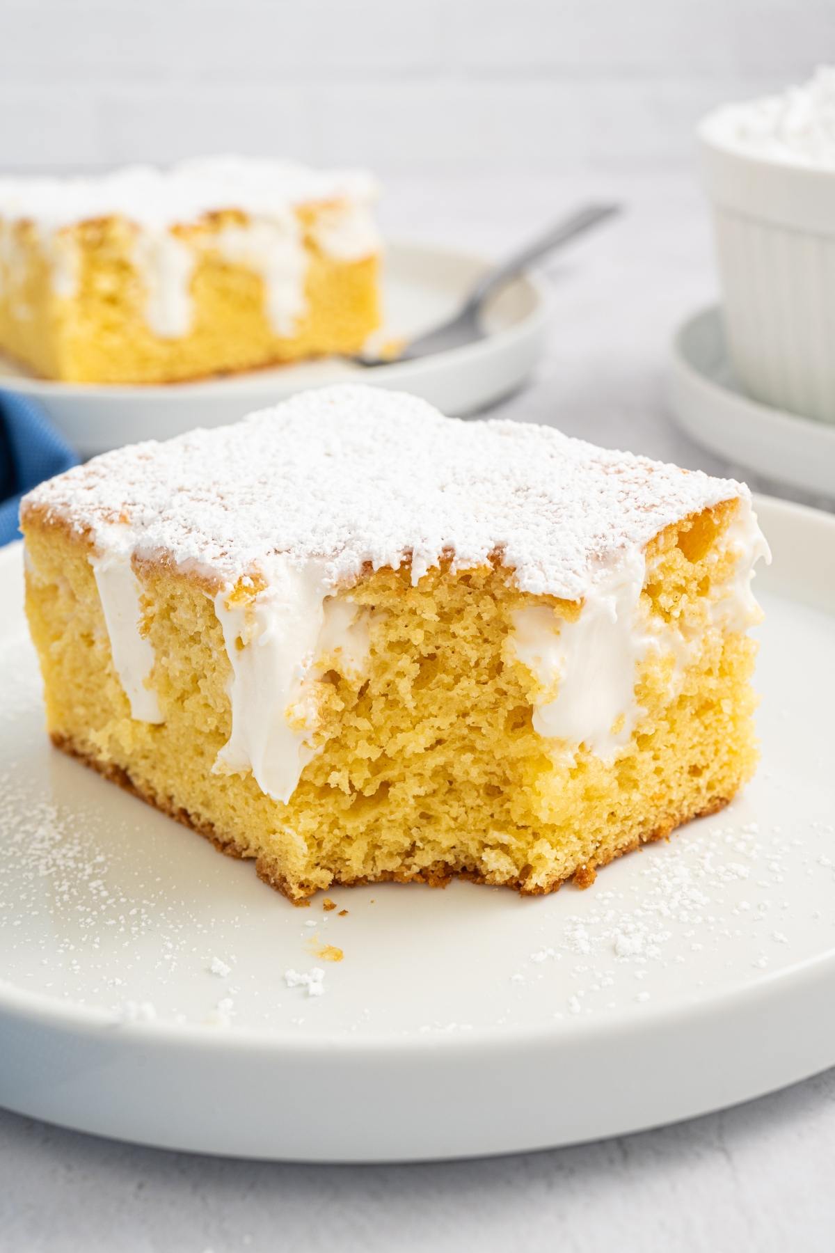 A square slice of yellow cake with white frosting and powdered sugar on a white plate.