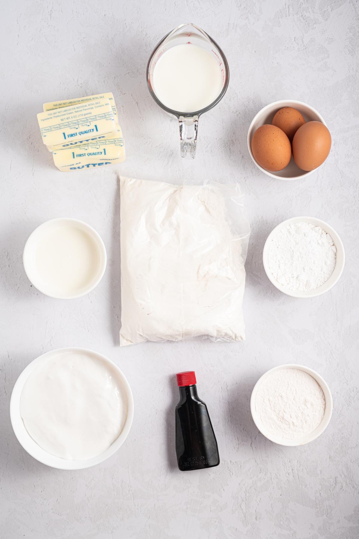 Baking ingredients: milk, eggs, butter, flour, sugar, vanilla extract, and bowls of other powders on a light surface.