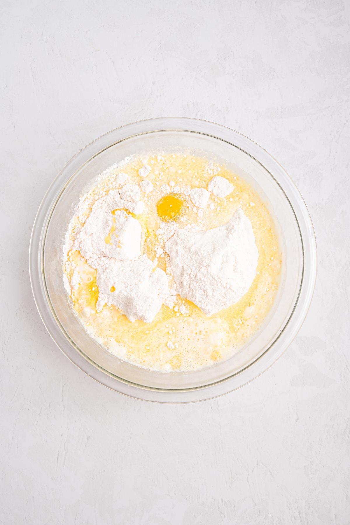 Clear glass bowl with flour, sugar, eggs, and wet ingredients on a light-colored surface, ready to be mixed.