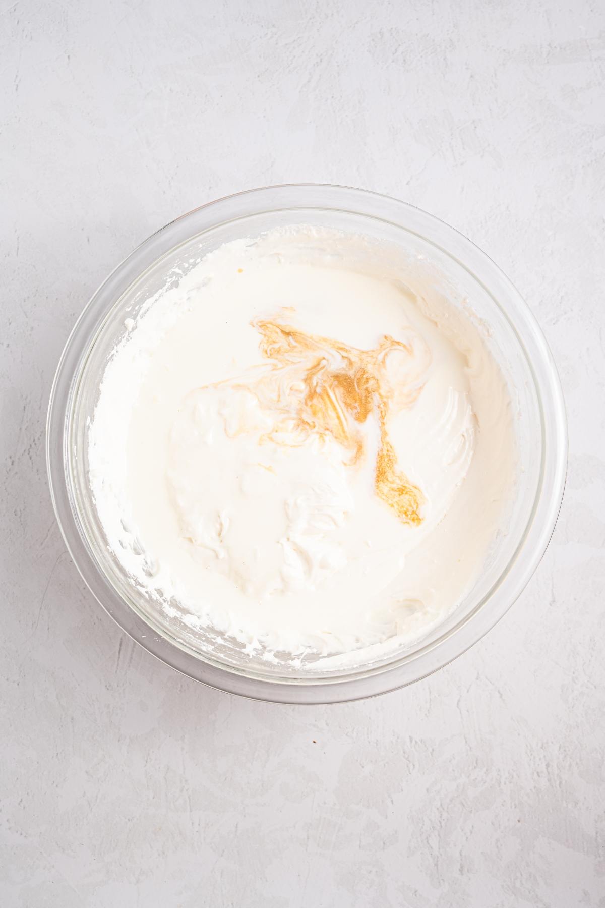 A glass bowl of whipped cream with vanilla extract being mixed in, on a light textured surface.