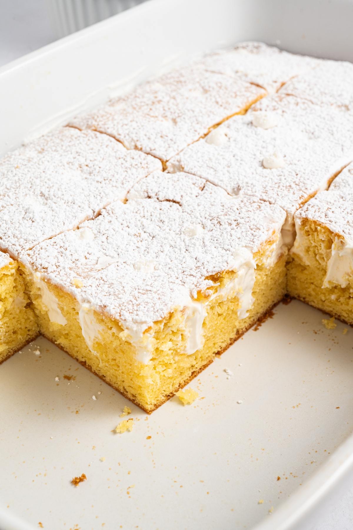 Sliced yellow cake with powdered sugar on top in a white baking dish, with a few pieces removed.
