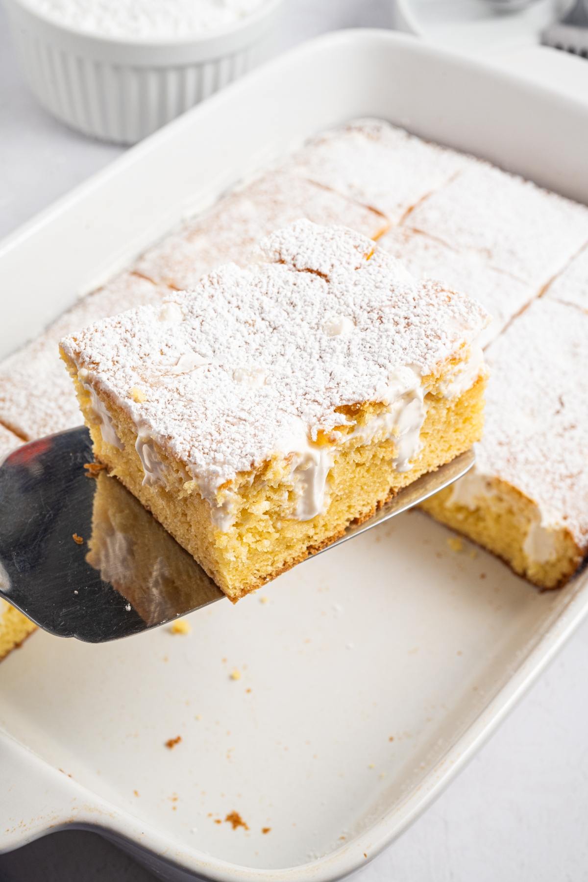 A slice of yellow cake with powdered sugar on top is being lifted from a baking dish.