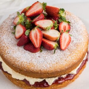 A classic Victoria Sandwich Cake featuring a light sponge layered with cream, jam, and fresh strawberries, finished with a dusting of powdered sugar on top.