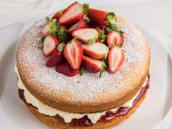 A classic Victoria Sandwich Cake featuring a light sponge layered with cream, jam, and fresh strawberries, finished with a dusting of powdered sugar on top.