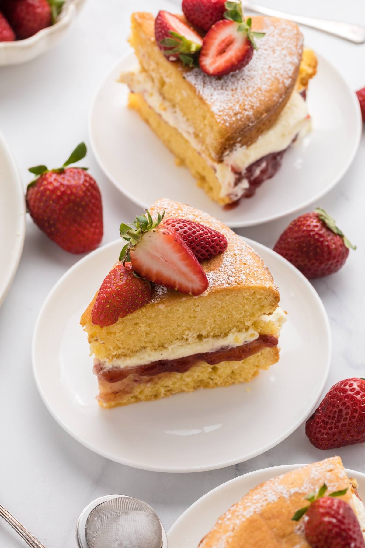 A slice of sponge cake with cream, jam, and strawberries on top, served on a white plate with fresh strawberries.