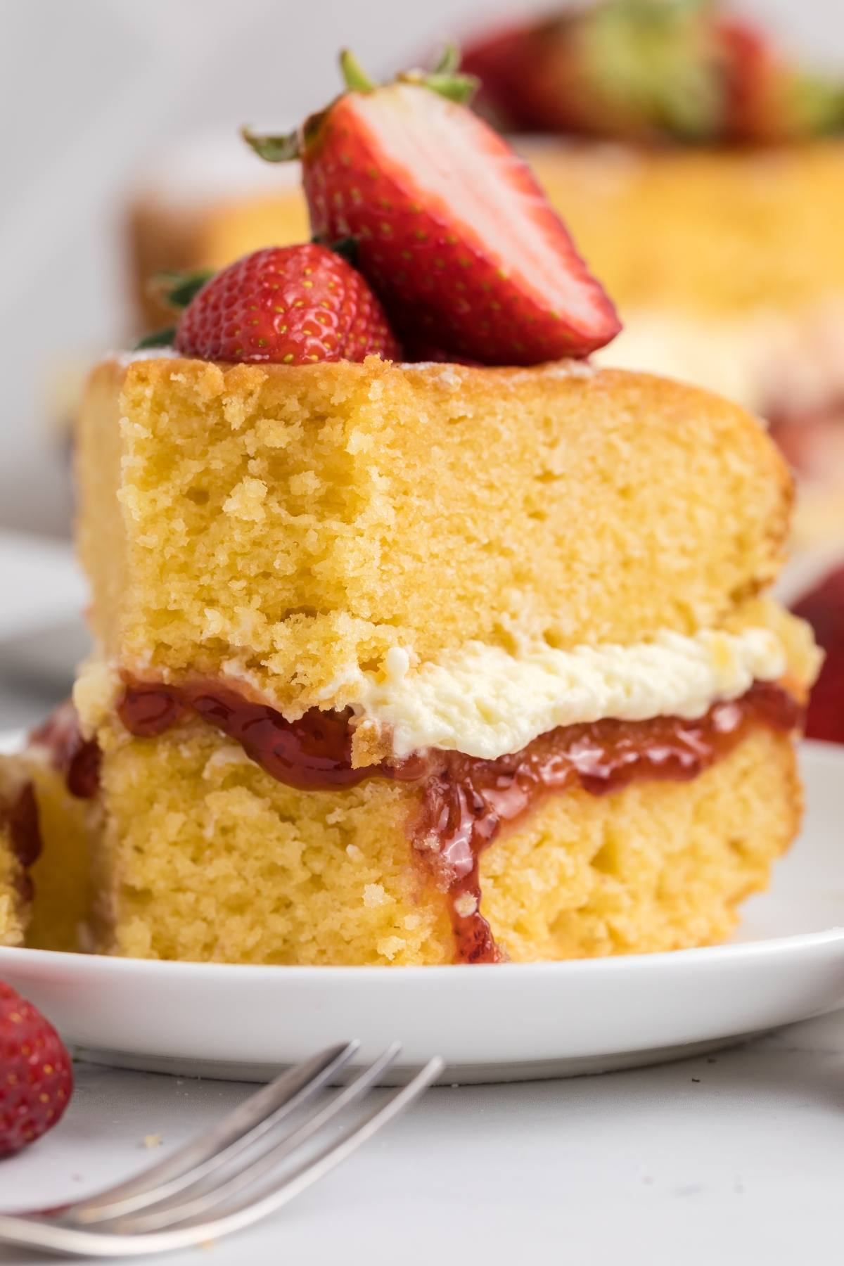 A slice of sponge cake with cream, strawberry jam, and fresh strawberries on top, served on a white plate.