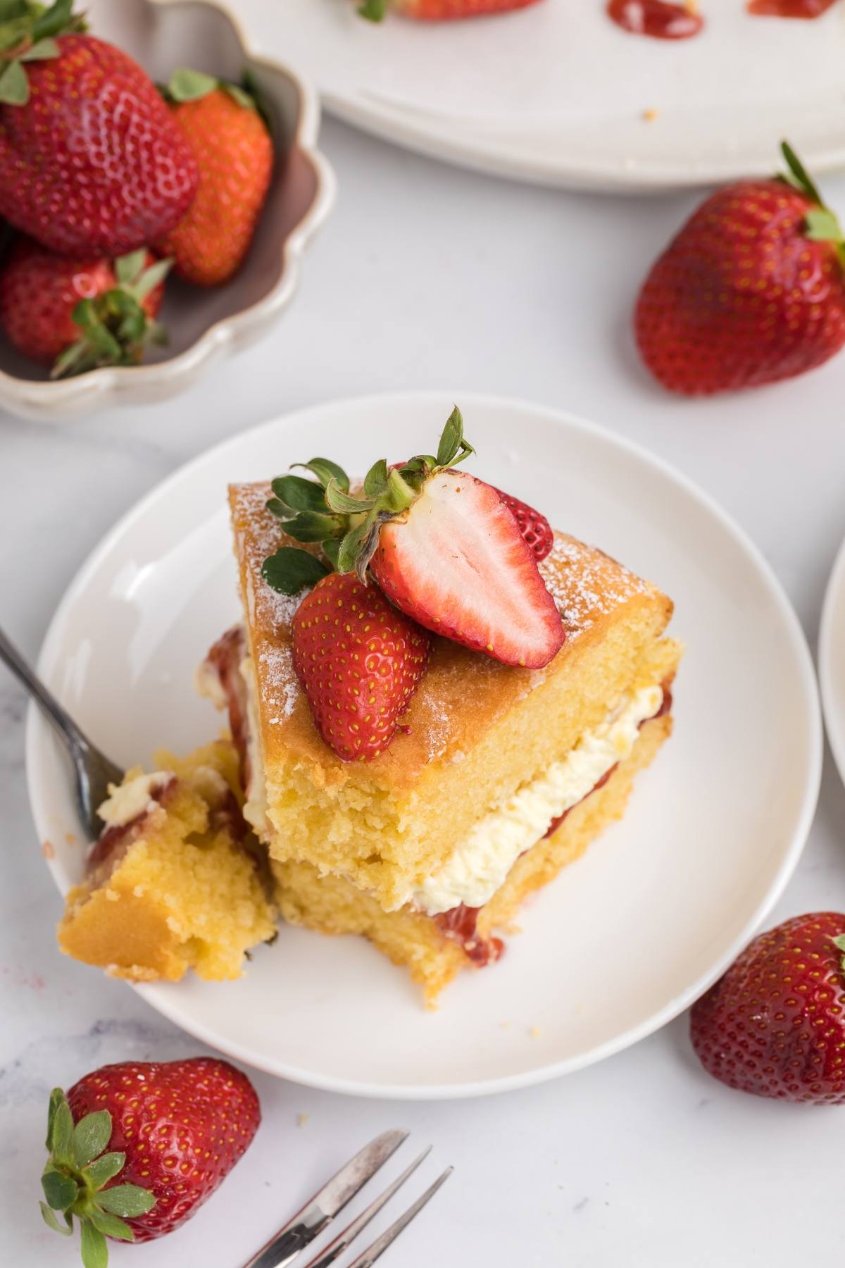 A slice of sponge cake with cream, jam, and strawberries on a white plate, surrounded by fresh strawberries.