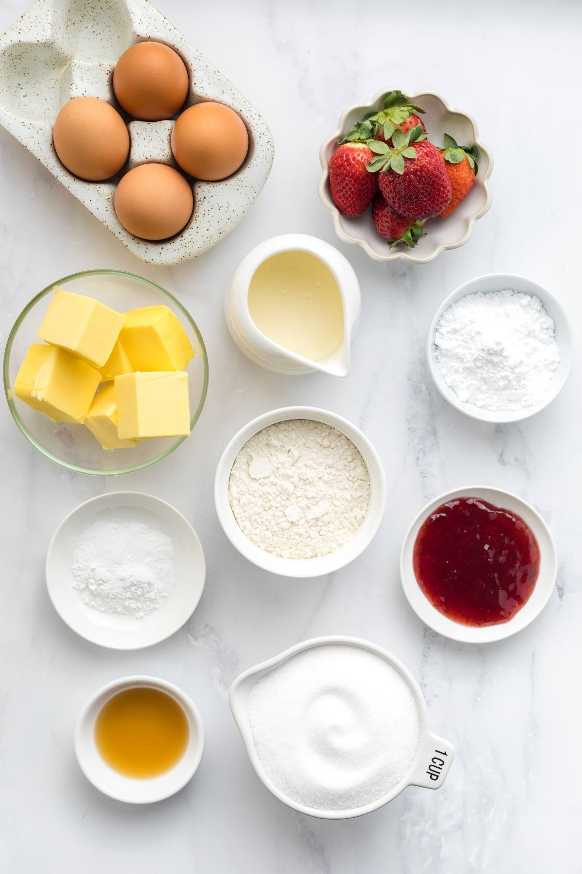 Top view of baking ingredients: eggs, butter, flour, sugar, strawberries, jam, vanilla, and powders in bowls.