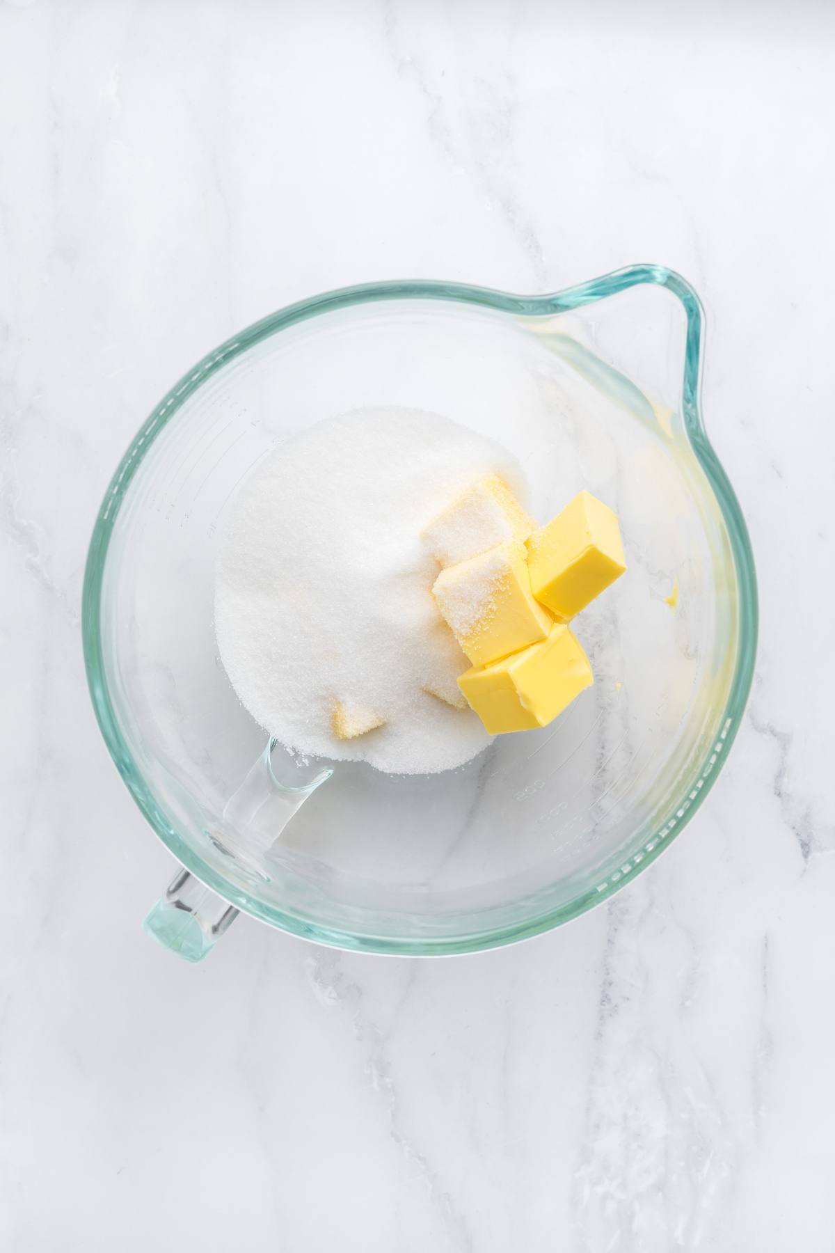 Glass mixing bowl with granulated sugar and cubes of butter on a white marble surface.