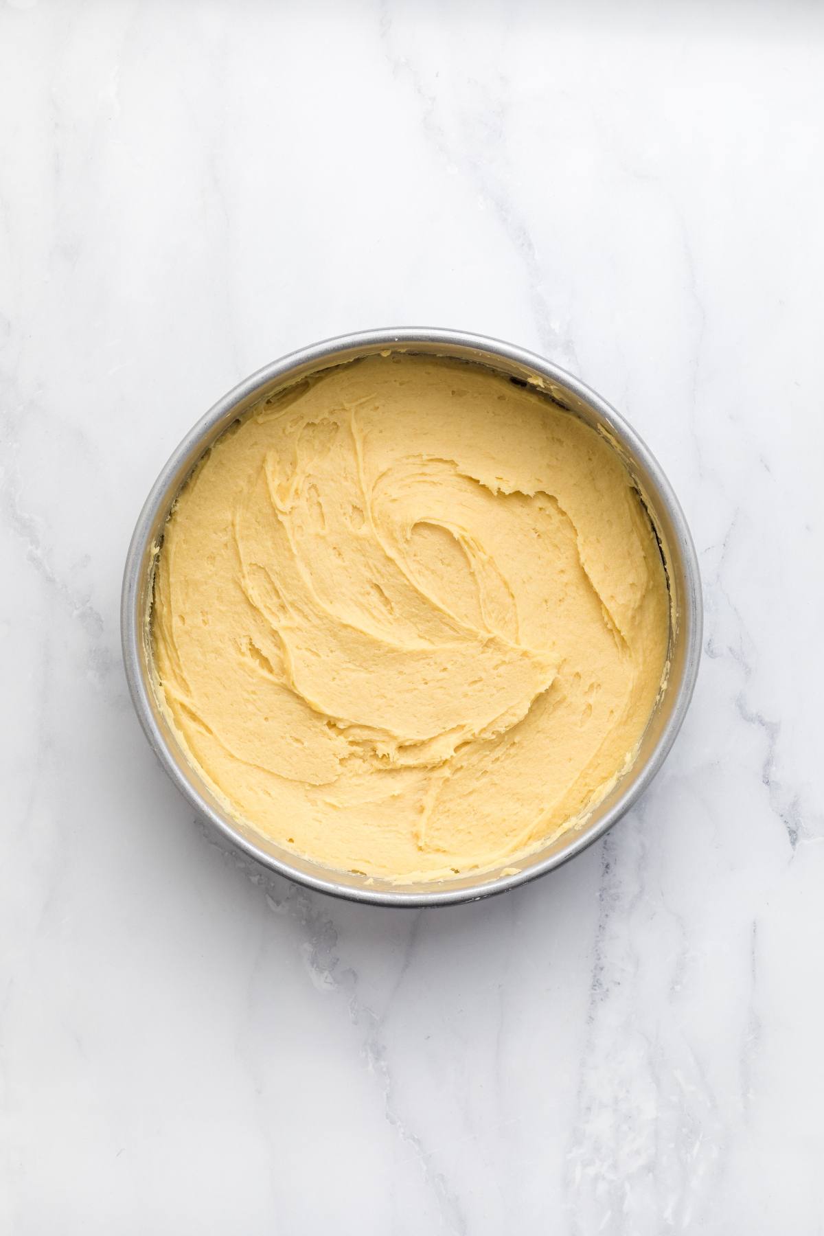 Round cake pan filled with smooth yellow cake batter on a white marble surface.