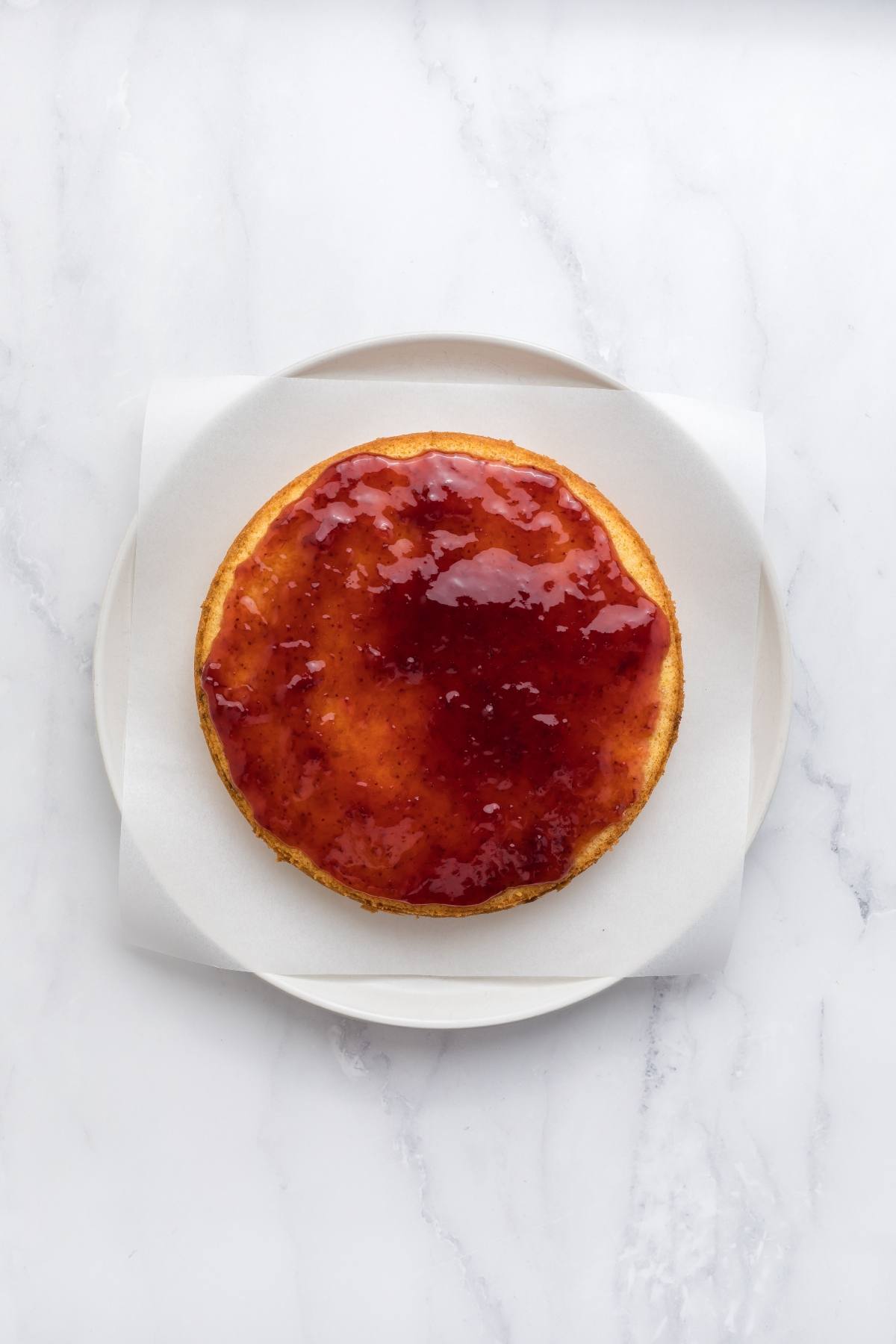 A round sponge cake topped with glossy red jam, placed on a white plate and parchment paper.