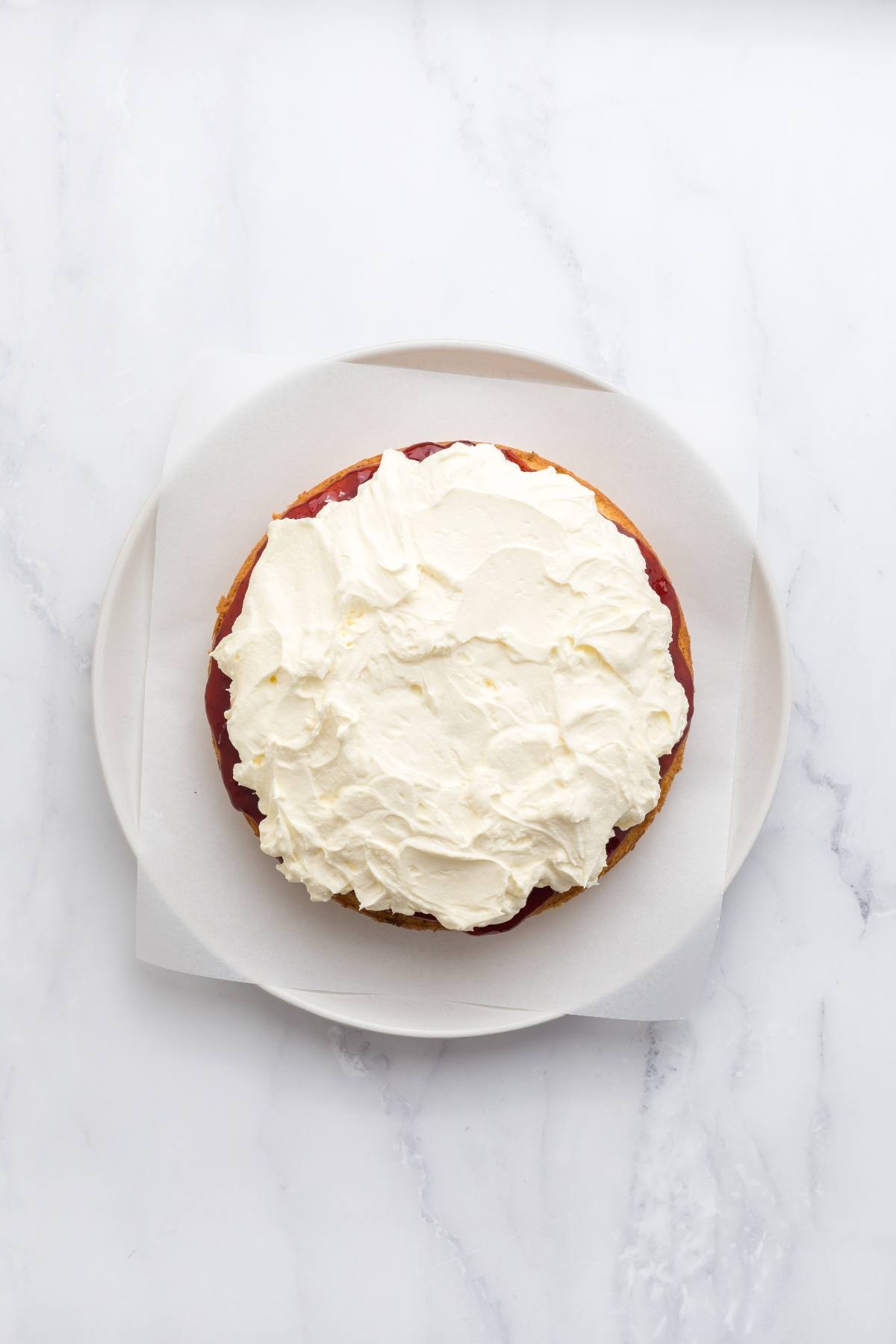 A round cake topped with a thick layer of whipped cream, placed on white parchment paper on a plate.