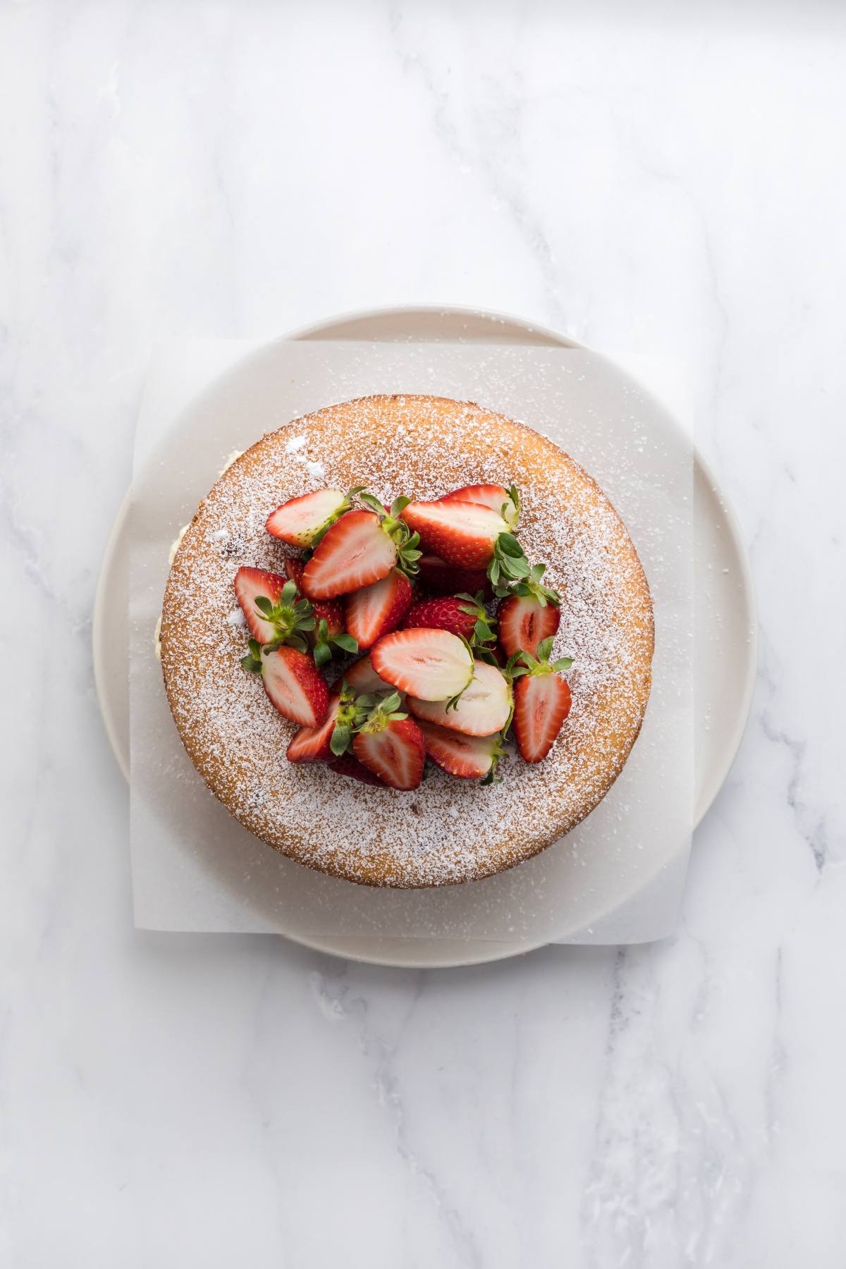 A round cake topped with sliced strawberries and powdered sugar on a white plate, seen from above.