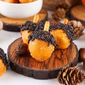 Mini acorn donut holes with chocolate sprinkles on wooden slices, surrounded by pinecones and acorns.