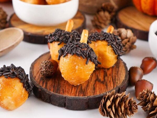 Mini acorn donut holes with chocolate sprinkles on wooden slices, surrounded by pinecones and acorns.