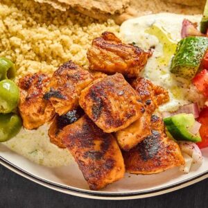 Aleppo Pepper Chicken Bites, couscous, olives, tzatziki, pita bread, and a cucumber-tomato salad on a plate.