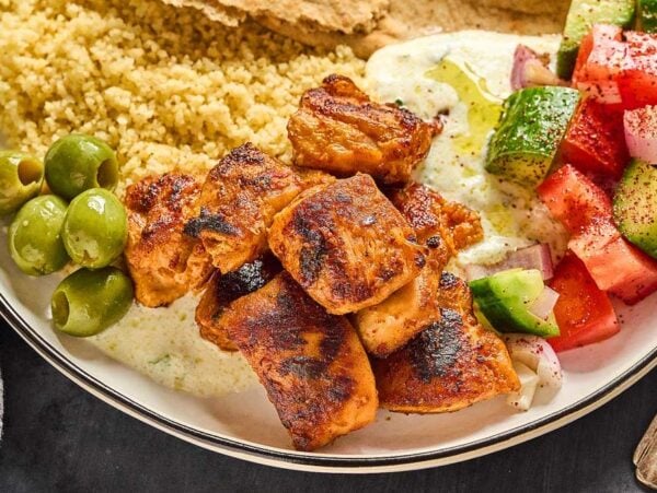 Aleppo Pepper Chicken Bites, couscous, olives, tzatziki, pita bread, and a cucumber-tomato salad on a plate.